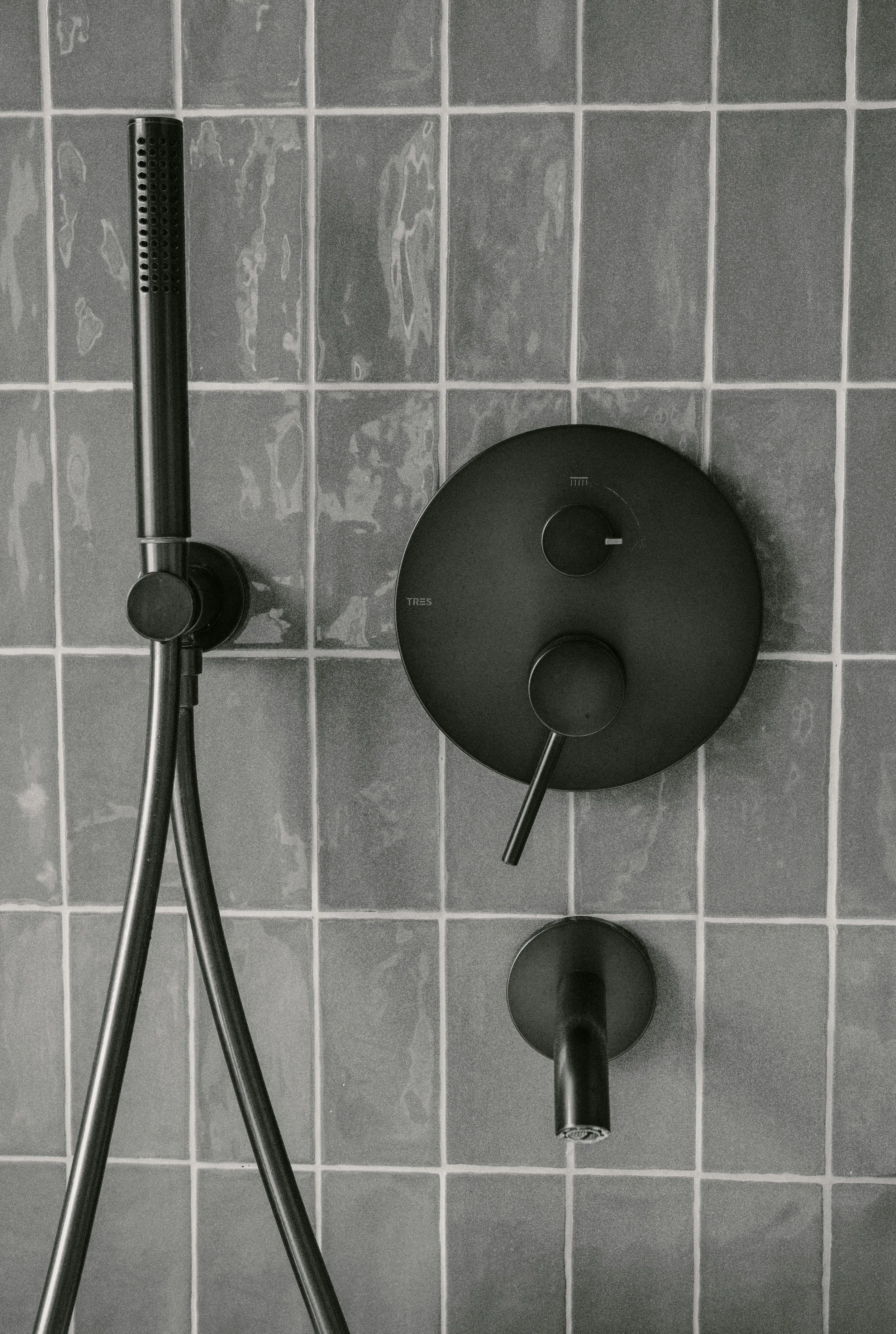 A shower area with gray tiled wall, a black shower head attached to a metal pole on the left, and a black round control panel with two dials on the right.