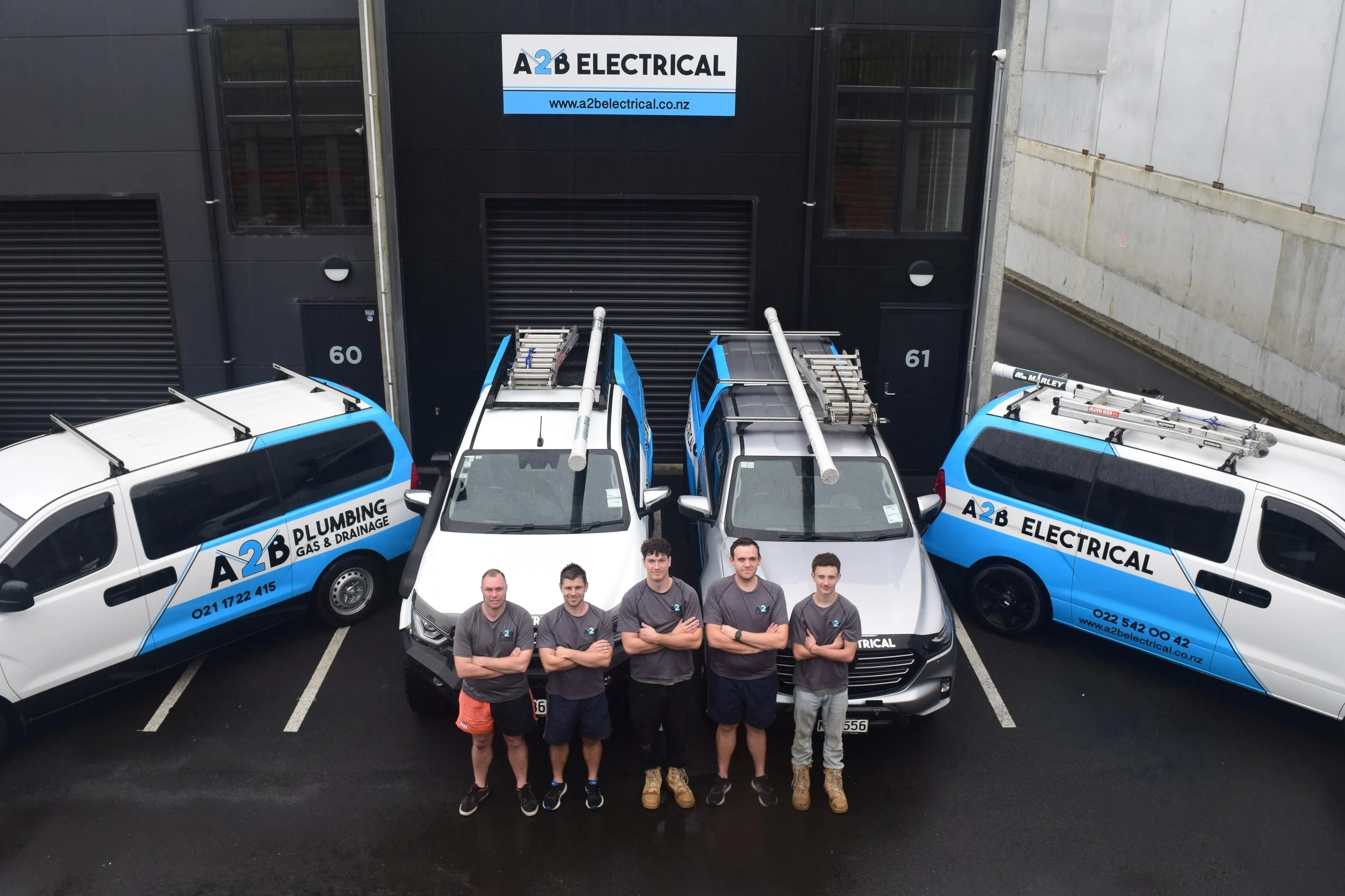 Construction team of six men standing in front of company vehicles and building with company sign, A2B Electrical.