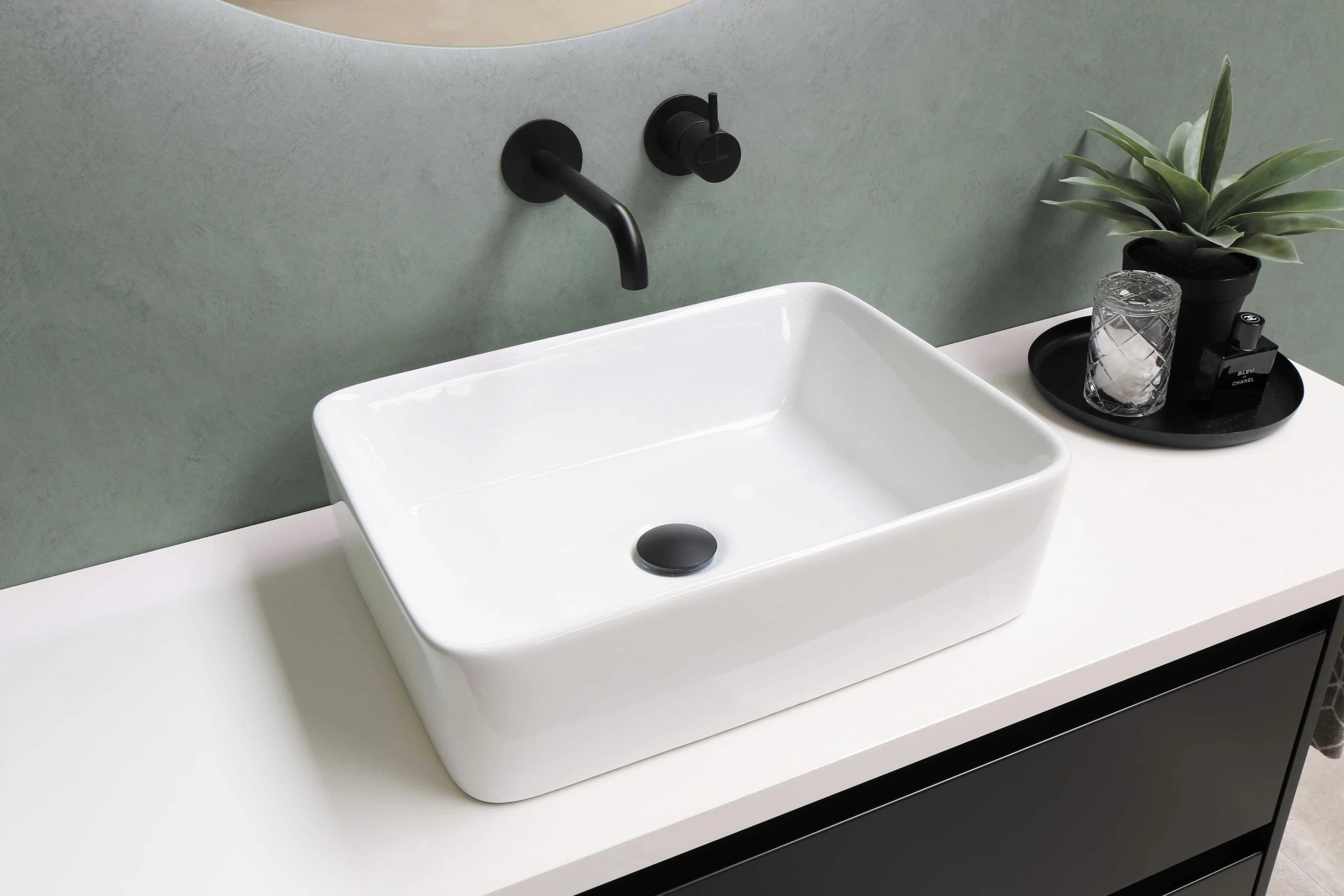 A white rectangular bathroom sink with a black drain stopper, mounted on a white countertop. Above it, a black wall-mounted faucet and handle. To the right, a black tray with a glass tumbler filled with ice, a potted plant with broad green leaves, and a bottle of Bleu de Chanel perfume. The background wall is a pale green color.