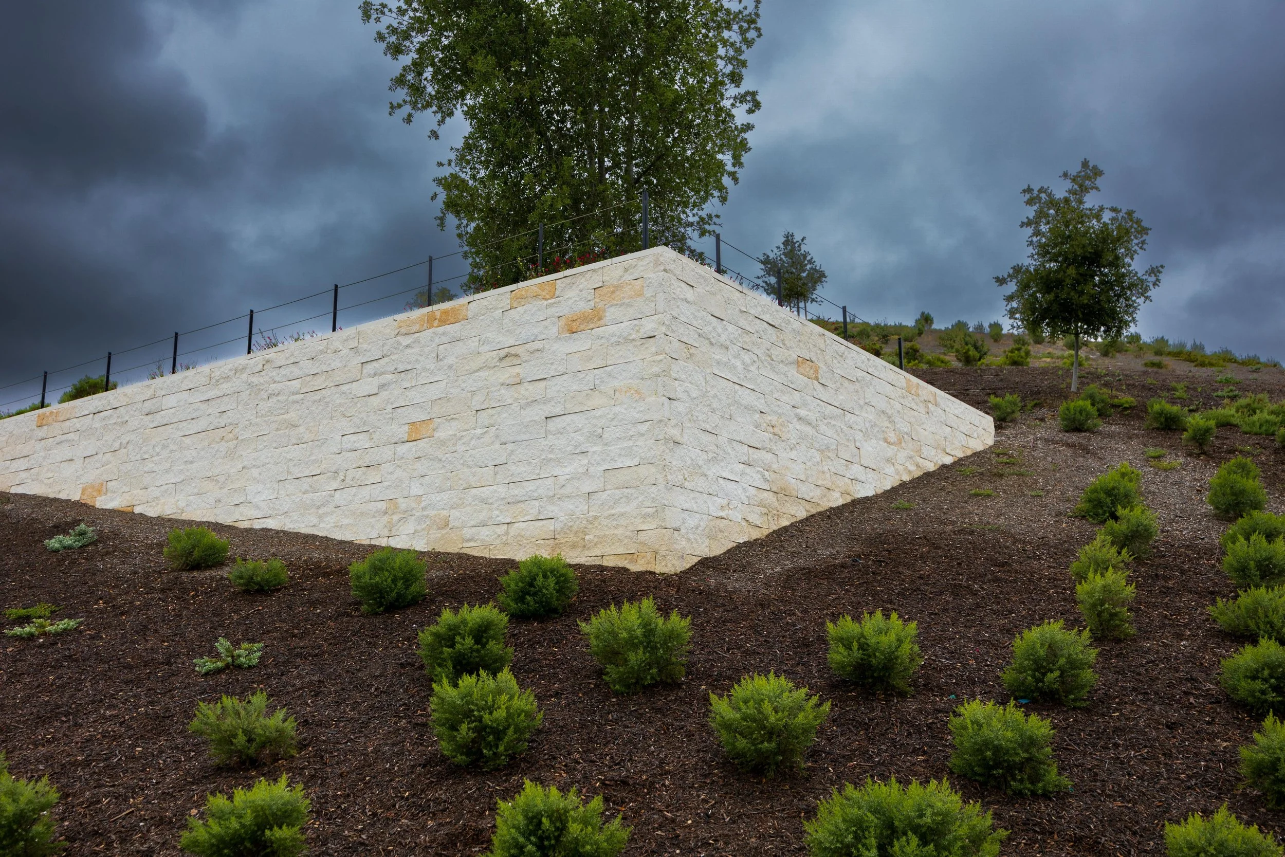 A stone retaining wall built of white and beige bricks with a black metal fence on top, extending along a landscaped hillside with small green bushes. Tall trees and dark stormy clouds are visible in the background.