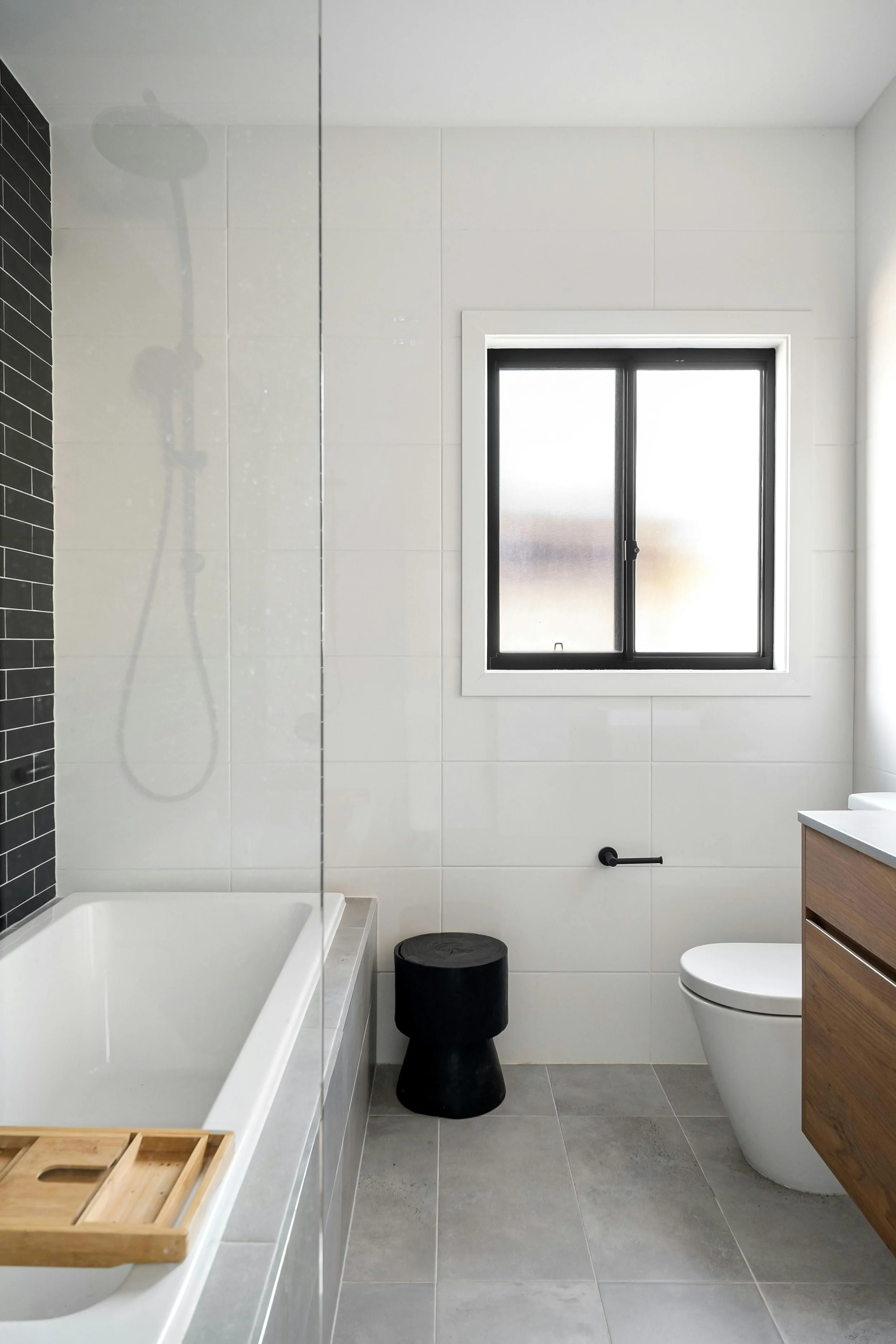 Modern bathroom with white tiled walls, a tub with a wooden tray, black stool, small black trash can, a window with frosted glass, and a wooden cabinet above the toilet.
