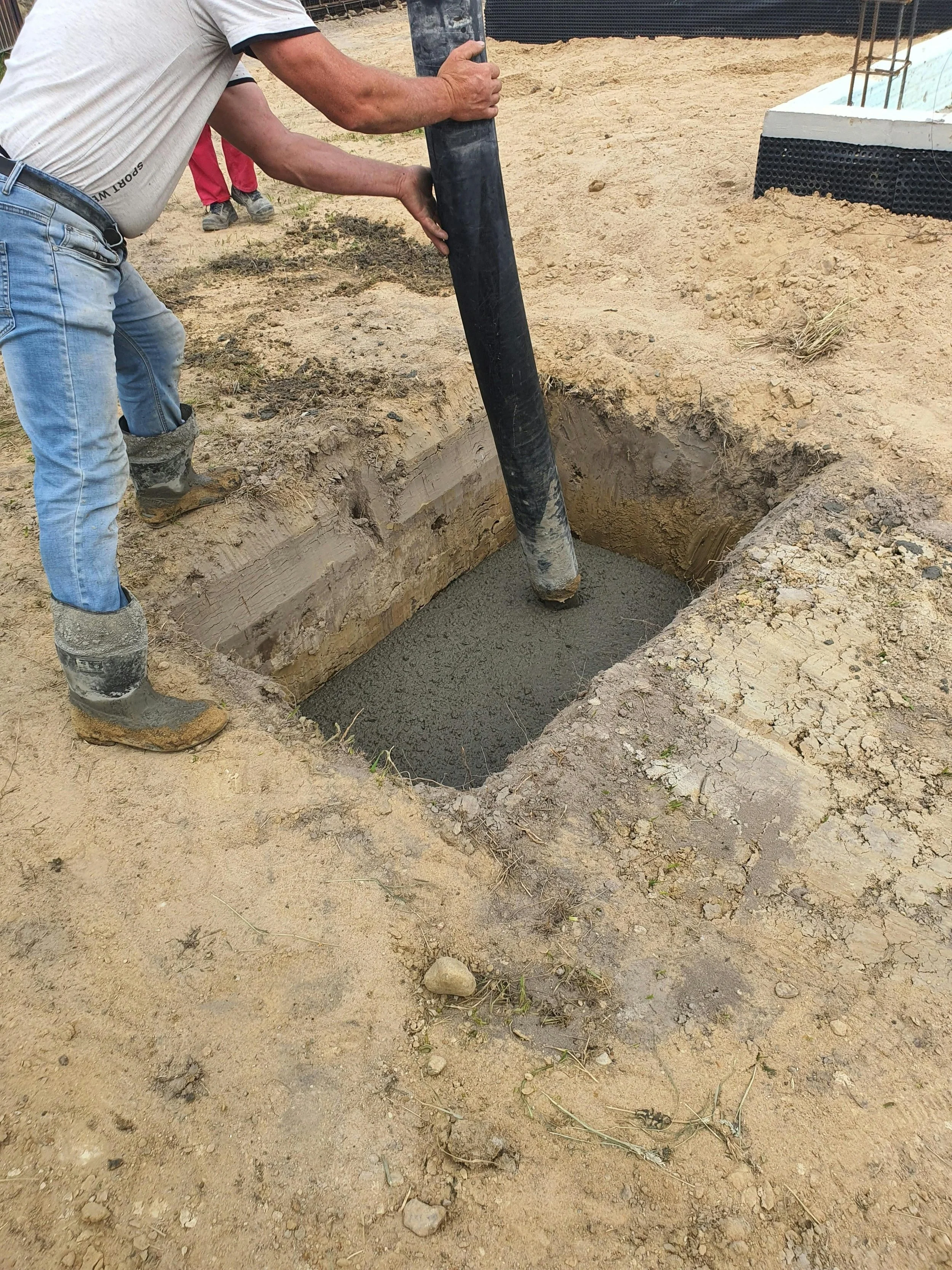 Person pouring wet concrete into a rectangular excavation at a construction site.