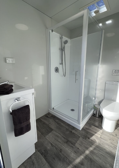 A clean, modern bathroom featuring a glass shower stall, a white toilet, a small white vanity with a folded towel on top, and a dark towel hanging on a towel ring. The flooring is grey wood-look vinyl, and a ceiling skylight provides natural light.