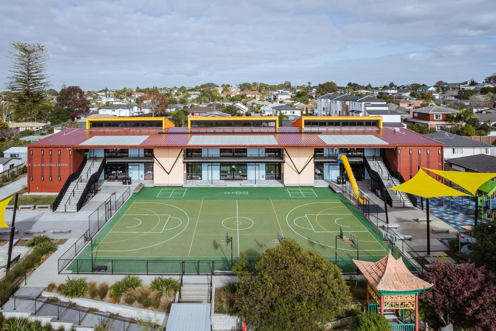Aerial view of a modern school building with a sports field, playground, and surrounding residential neighborhood.