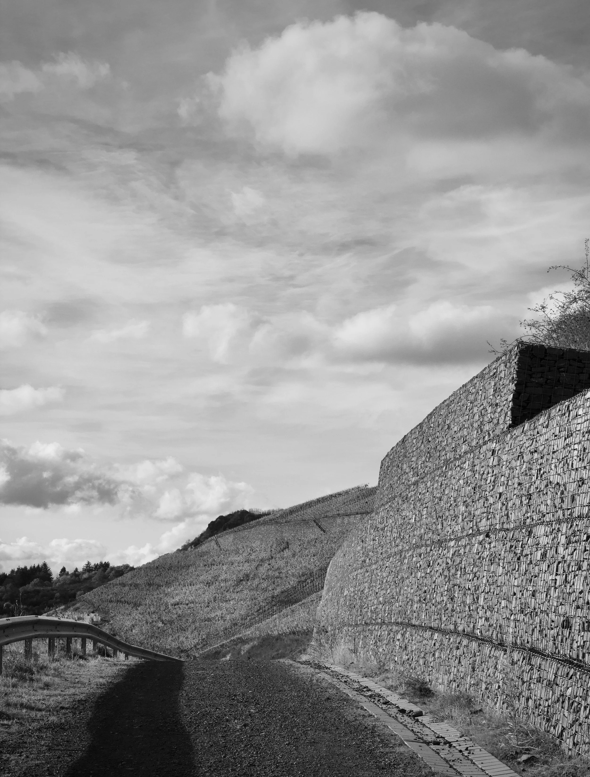 A winding country road with a guardrail on the left, a hillside with trees in the background, and a large valley with a stone retaining wall on the right under a cloudy sky.
