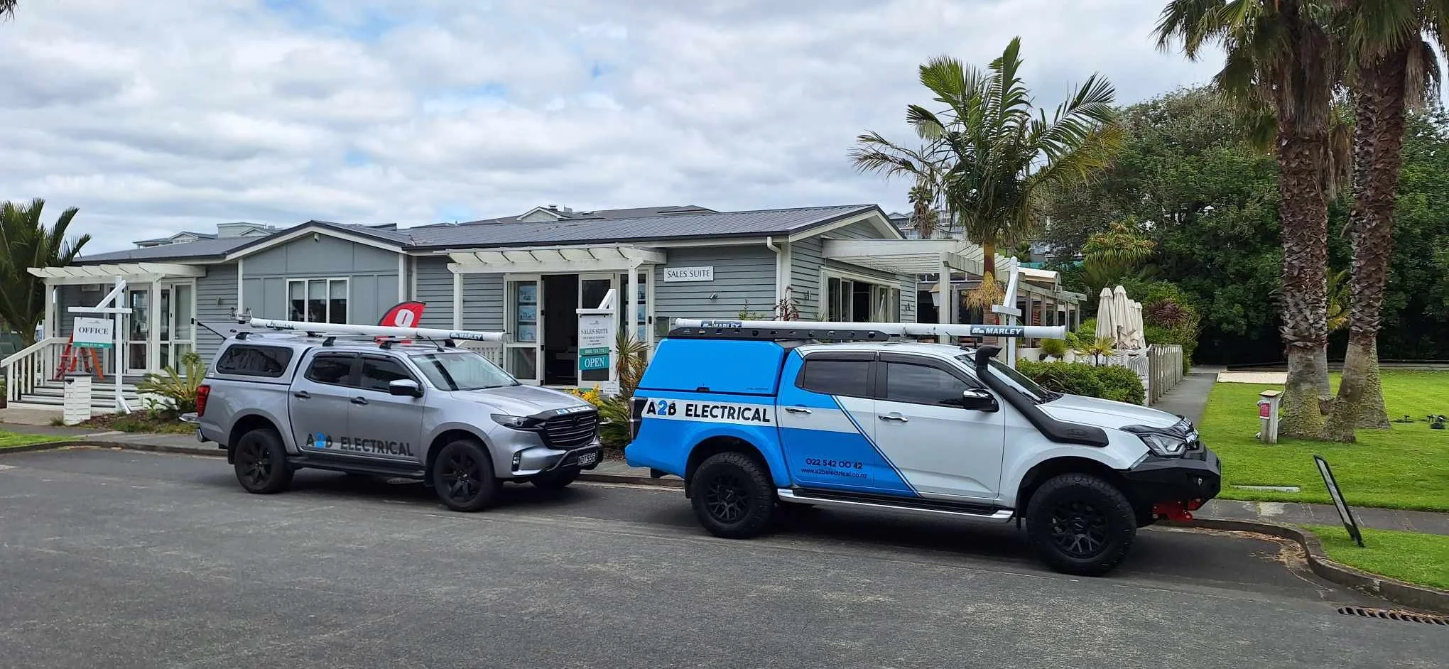 Two electrical service trucks parked in front of a light blue building with signs indicating an office and sales suite. There are palm trees and a grassy area nearby.