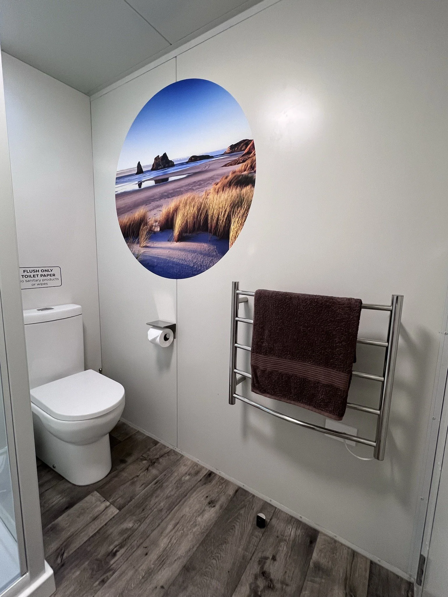 Public restroom with a toilet, a wall-mounted toilet paper dispenser, a towel rack with a maroon towel, and a large round wall art depicting a beach scene with rocks and dunes.