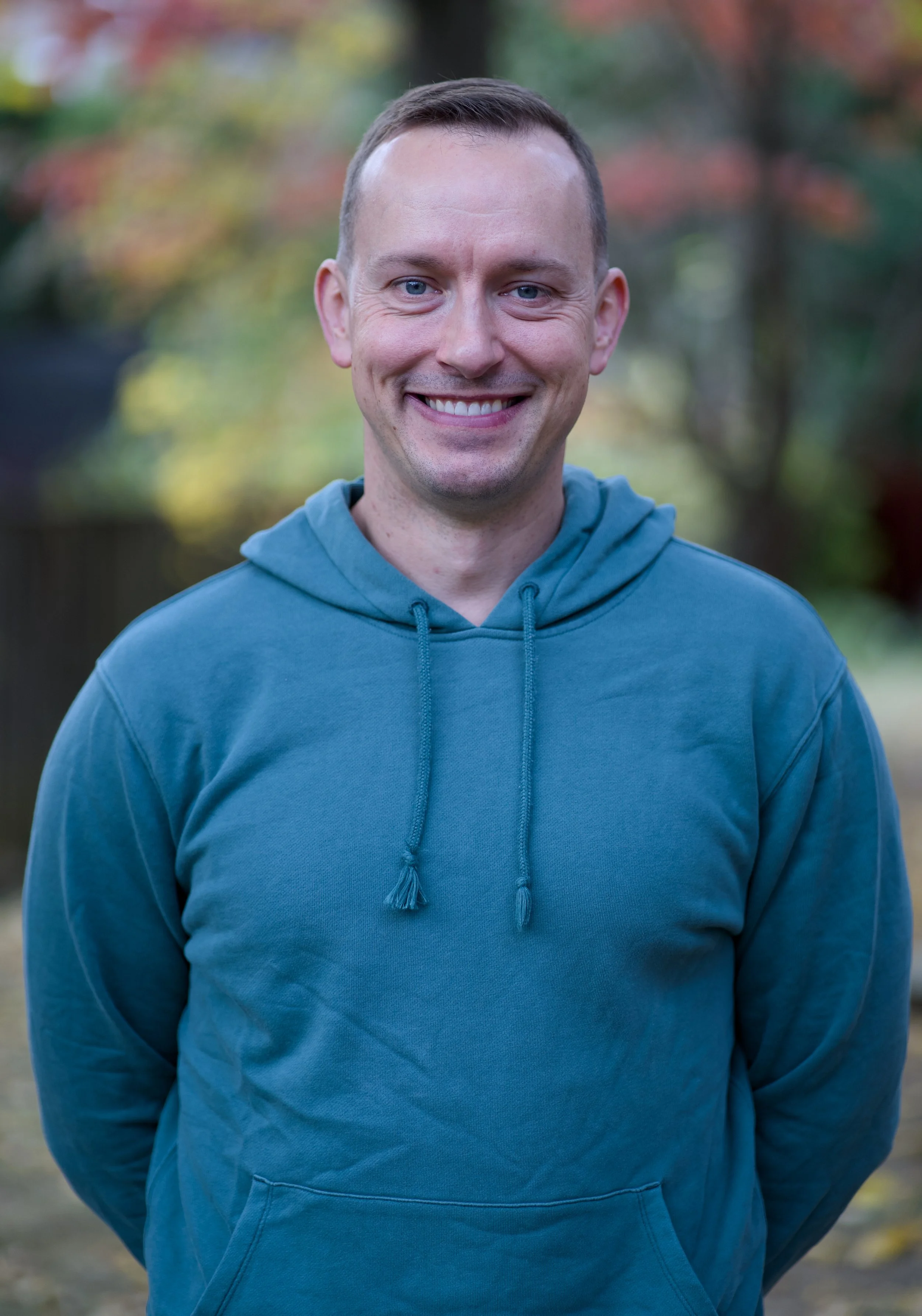 A smiling man with blue eyes and short brown hair wearing a blue hoodie, standing outdoors with blurred trees in fall colors in the background.