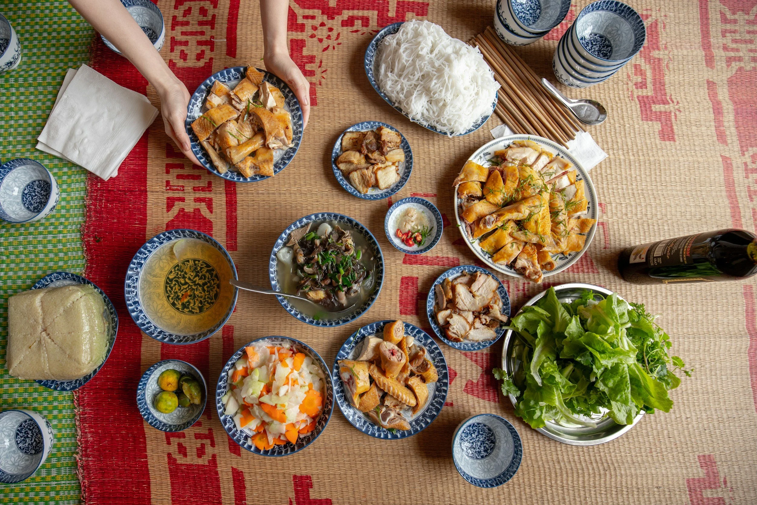 A top-down view of a traditional Asian meal spread on a mat, including bowls of rice noodles, various cooked meats, vegetables, and a leafy green salad, along with a loaf of bread, small pickles, condiments, glasses, and a bottle of wine.