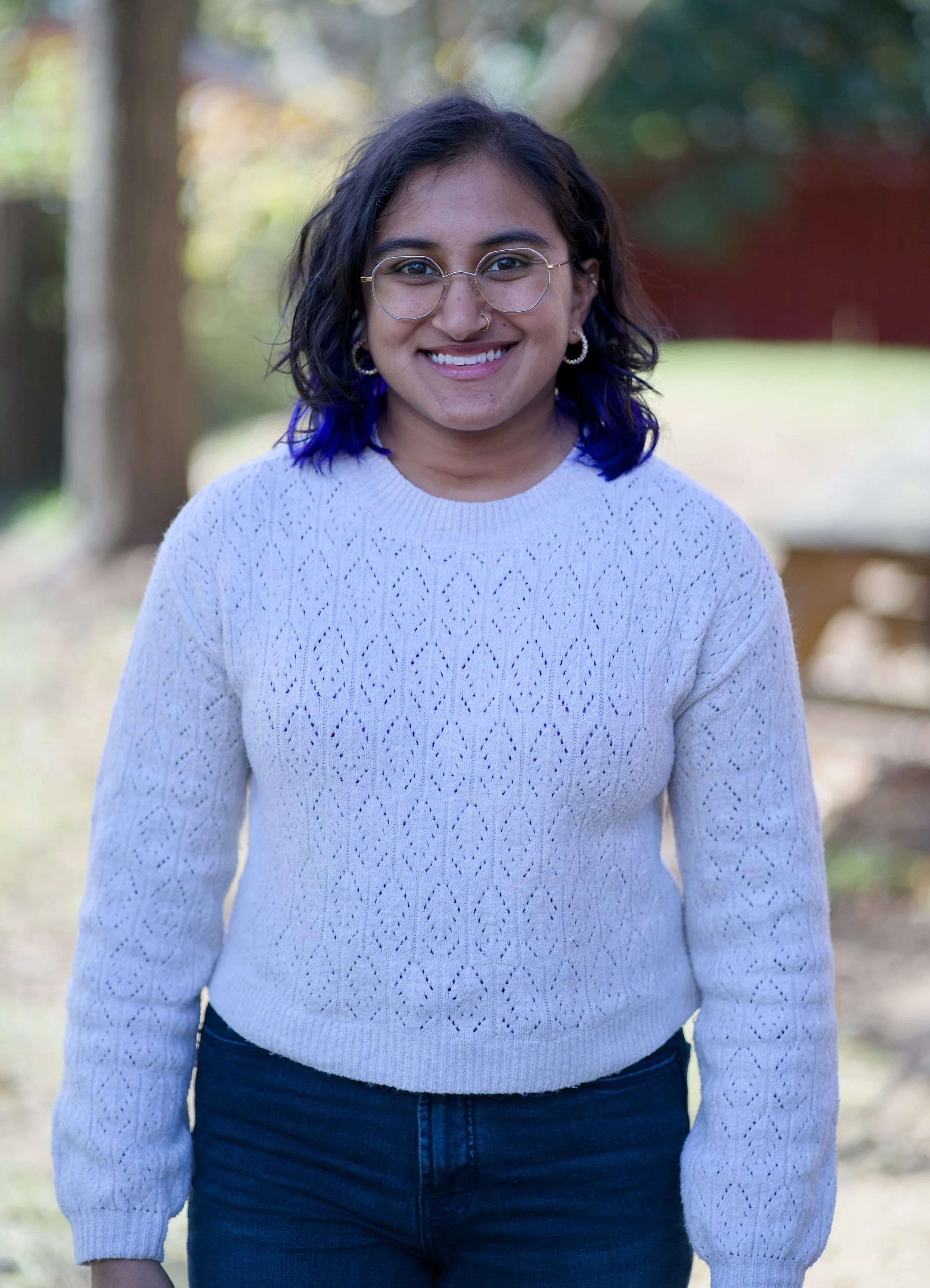 A young woman with glasses and a nose ring smiling outdoors, wearing a white sweater with a cable knit pattern and dark pants, with a blurred natural background of trees.