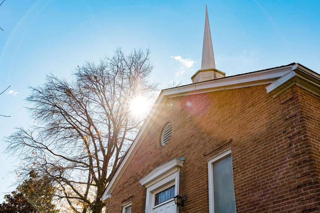 A brick church with a tall white steeple, a window, and a tree with bare branches in front, with sunlight streaming through the branches on a clear day.