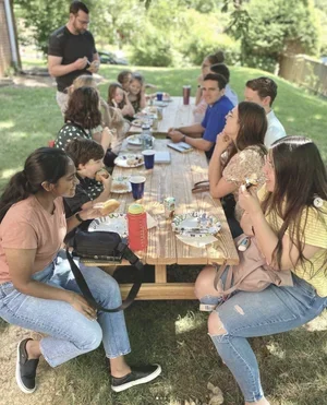 A group of young adults and some children gathered around a picnic table outdoors, having a meal together.