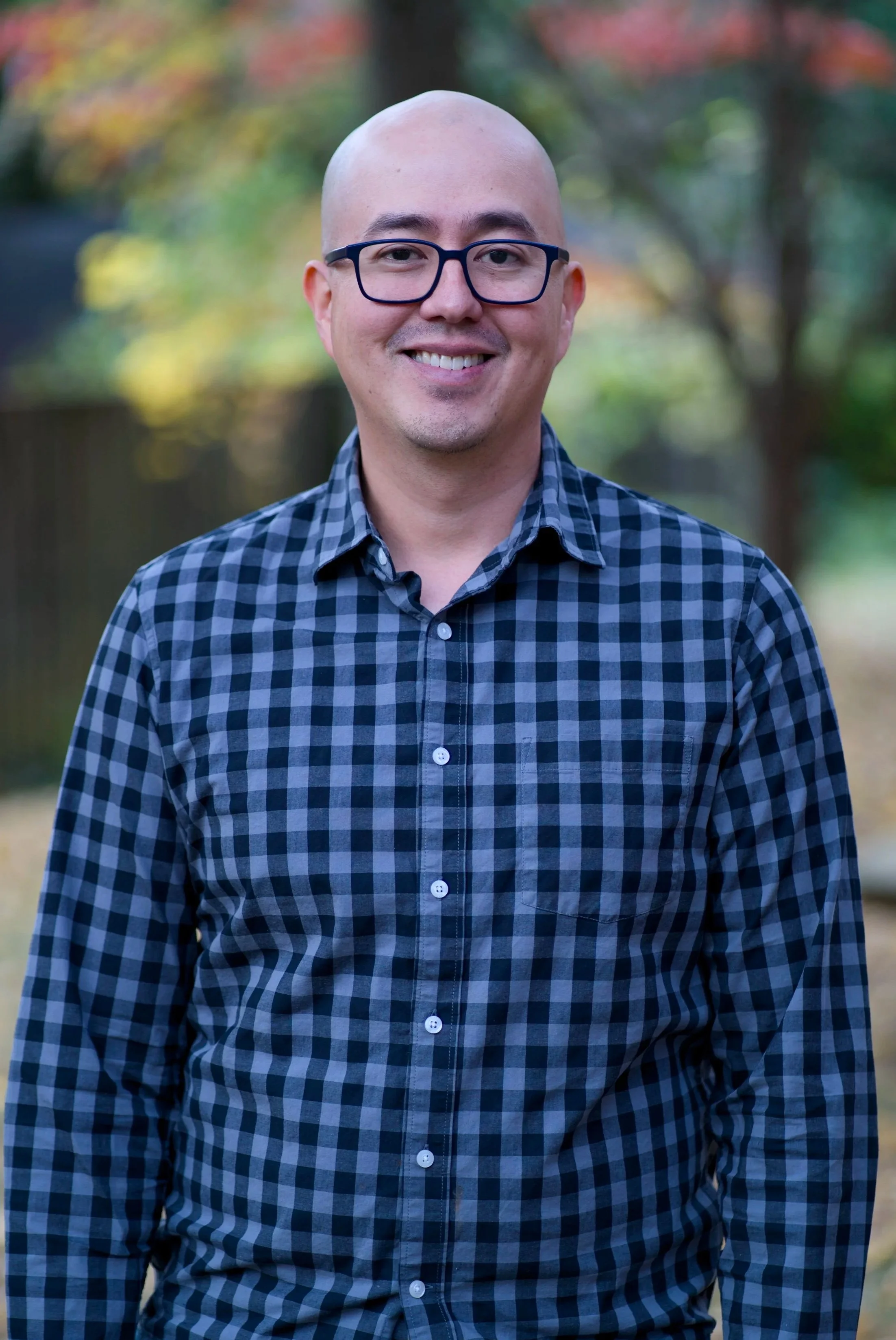 Portrait of a smiling man with glasses, wearing a checkered shirt, standing outdoors with blurred background of trees and foliage.