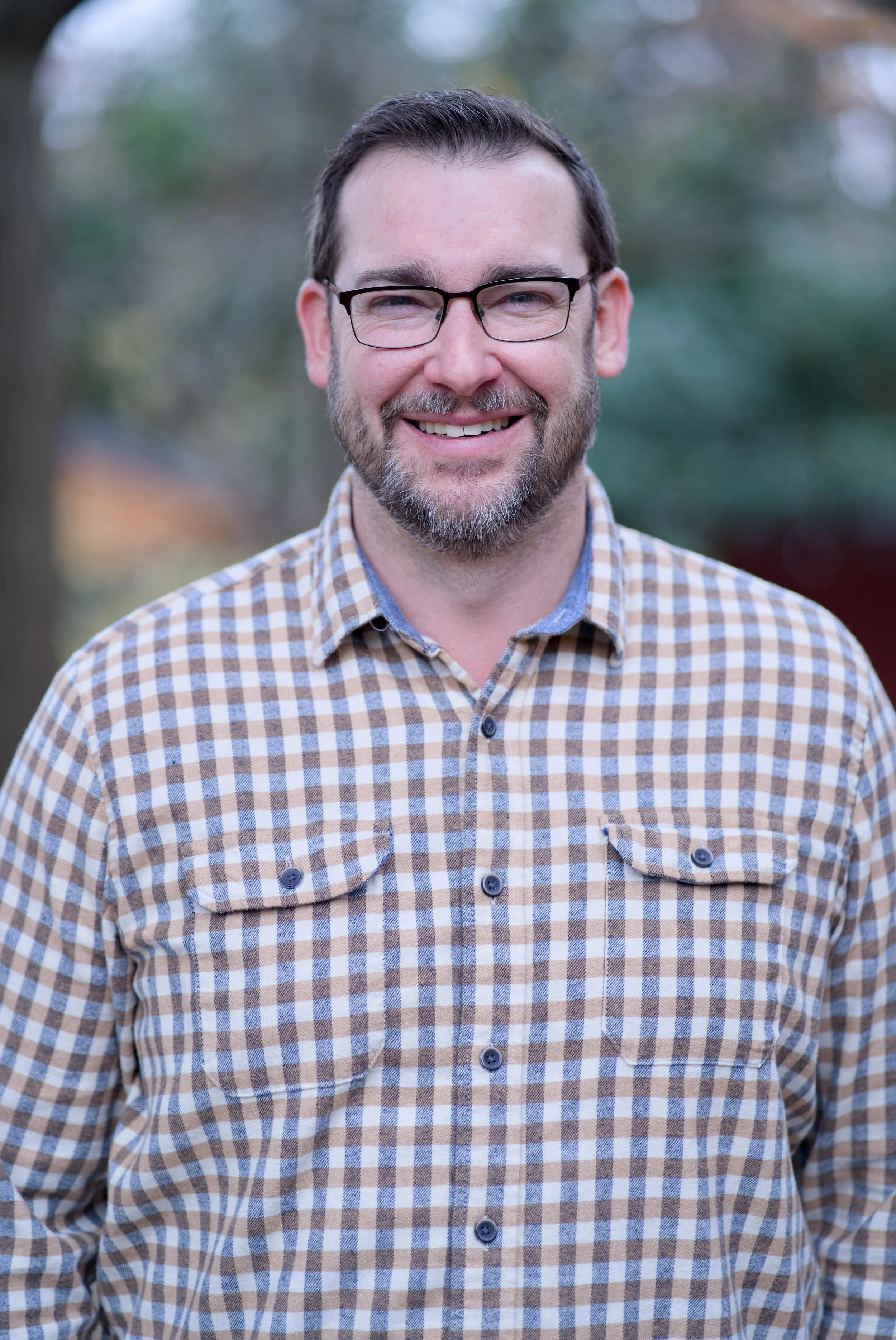 A smiling man with glasses, a beard, and short hair, wearing a beige and blue checkered shirt, standing outdoors with blurred trees in the background.
