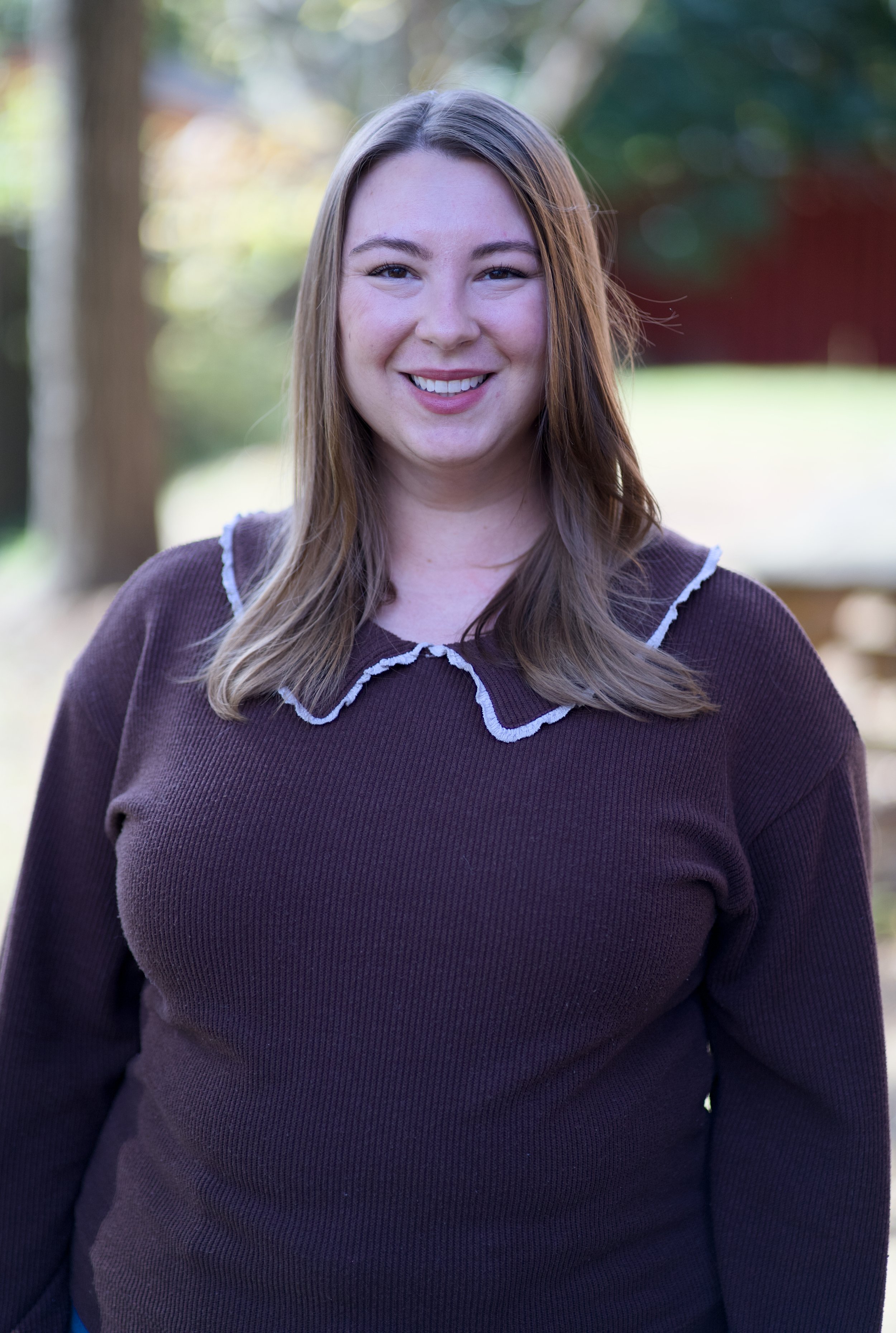 A young woman with long, light brown hair smiling outdoors, wearing a brown sweater with white trim on the collar, blurred trees and grass in the background.
