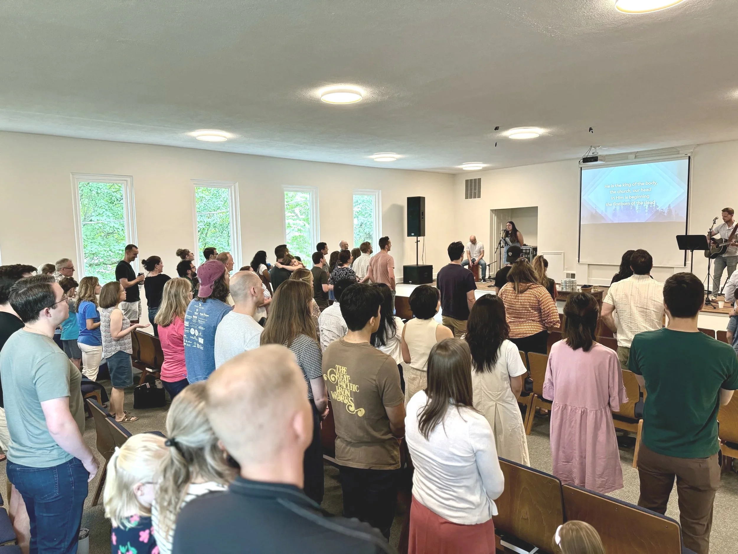 A large group of people attending indoor Christian worship service, some standing and some sitting, with a man playing guitar and a woman singing at the front, a large speaker, a projector screen displaying a hymn, and windows letting in natural light.