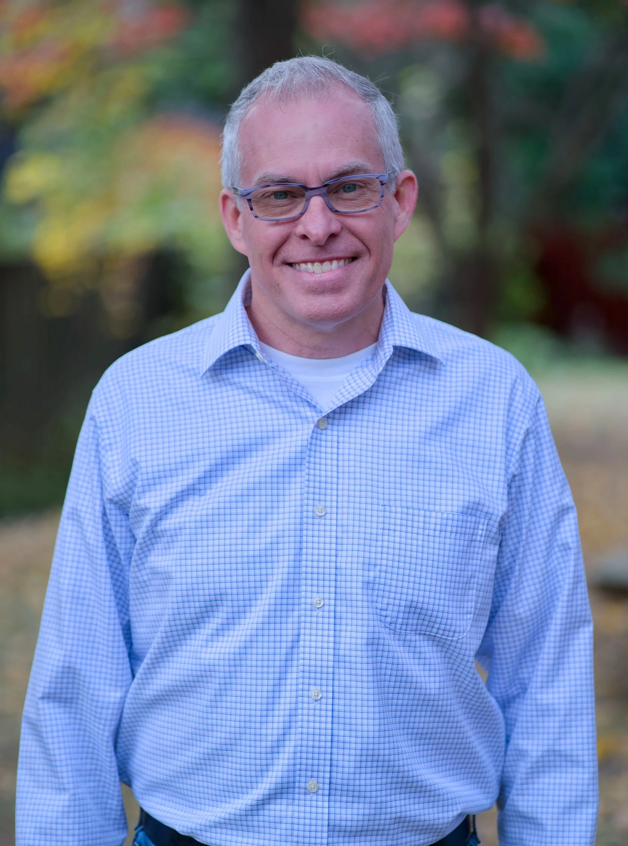 Portrait of a smiling middle-aged man with gray hair, wearing glasses and a blue checked shirt, standing outdoors with blurred colorful trees in the background.