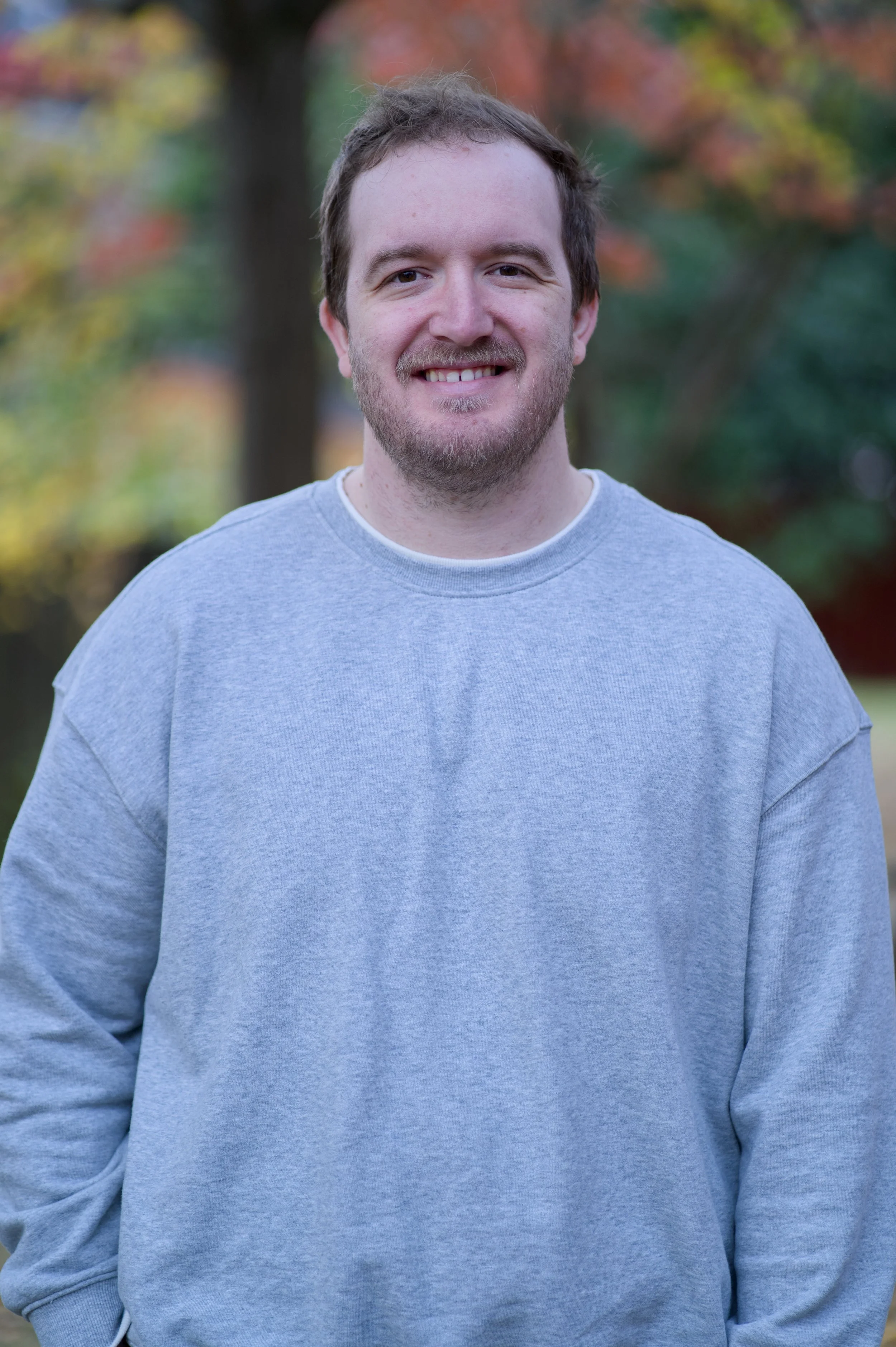 A young man with light skin, short brown hair, and a light beard, smiling while standing outdoors with blurred autumn-colored trees in the background, wearing a light gray sweatshirt.