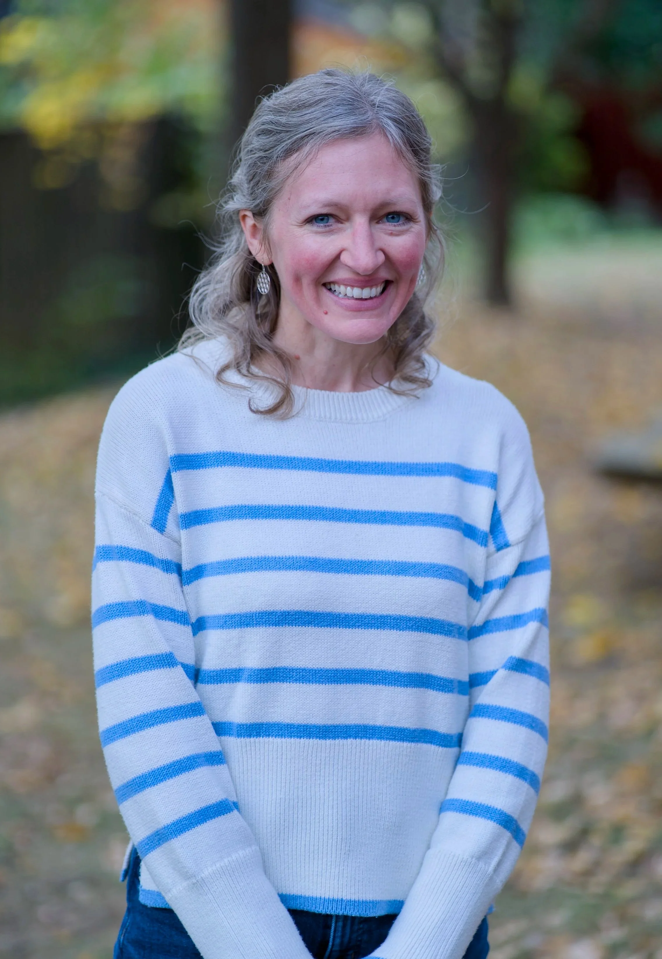 A woman with gray hair, blue eyes, and earrings smiling outdoors during fall, wearing a white sweater with blue stripes.