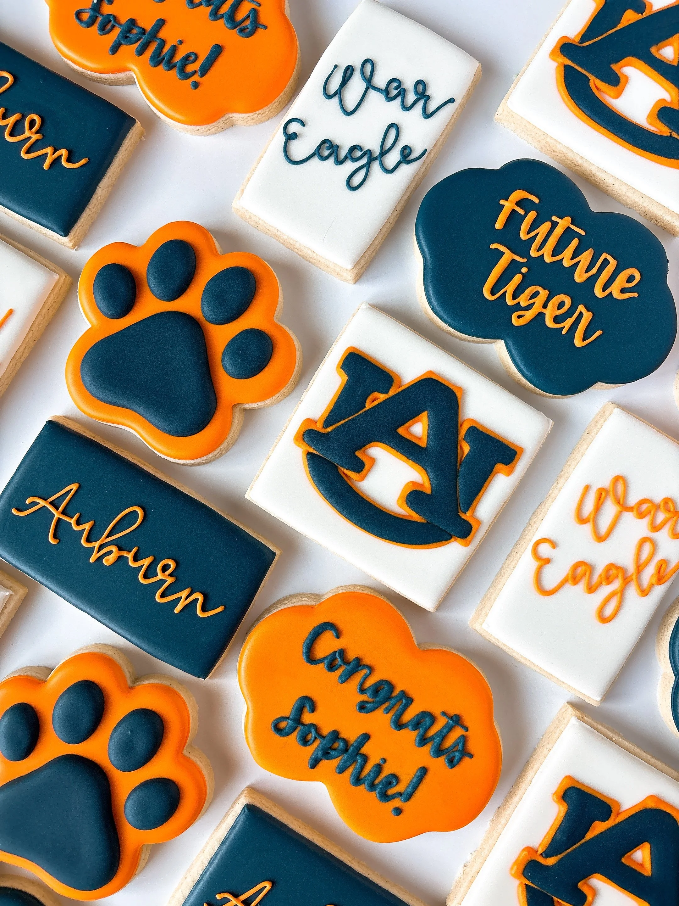 Decorative cookies decorated with Alabama-themed colors and text, including phrases like 'War Eagle' and 'Go Tigers', and featuring paw prints and university logos in navy blue, orange, and white.