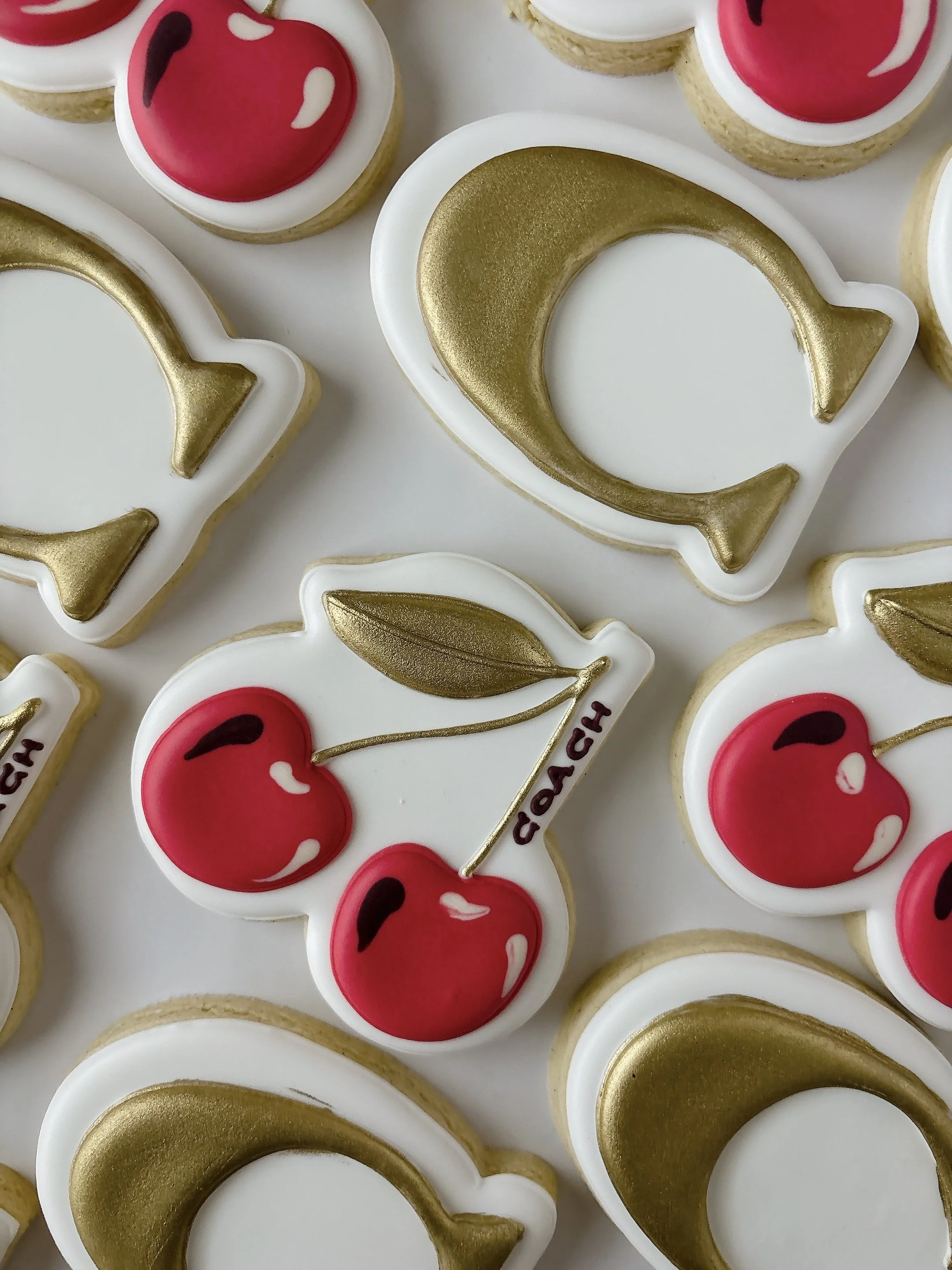 Arranged cookies decorated with red cherries and gold Coach logo, with some cookies featuring white outlines.