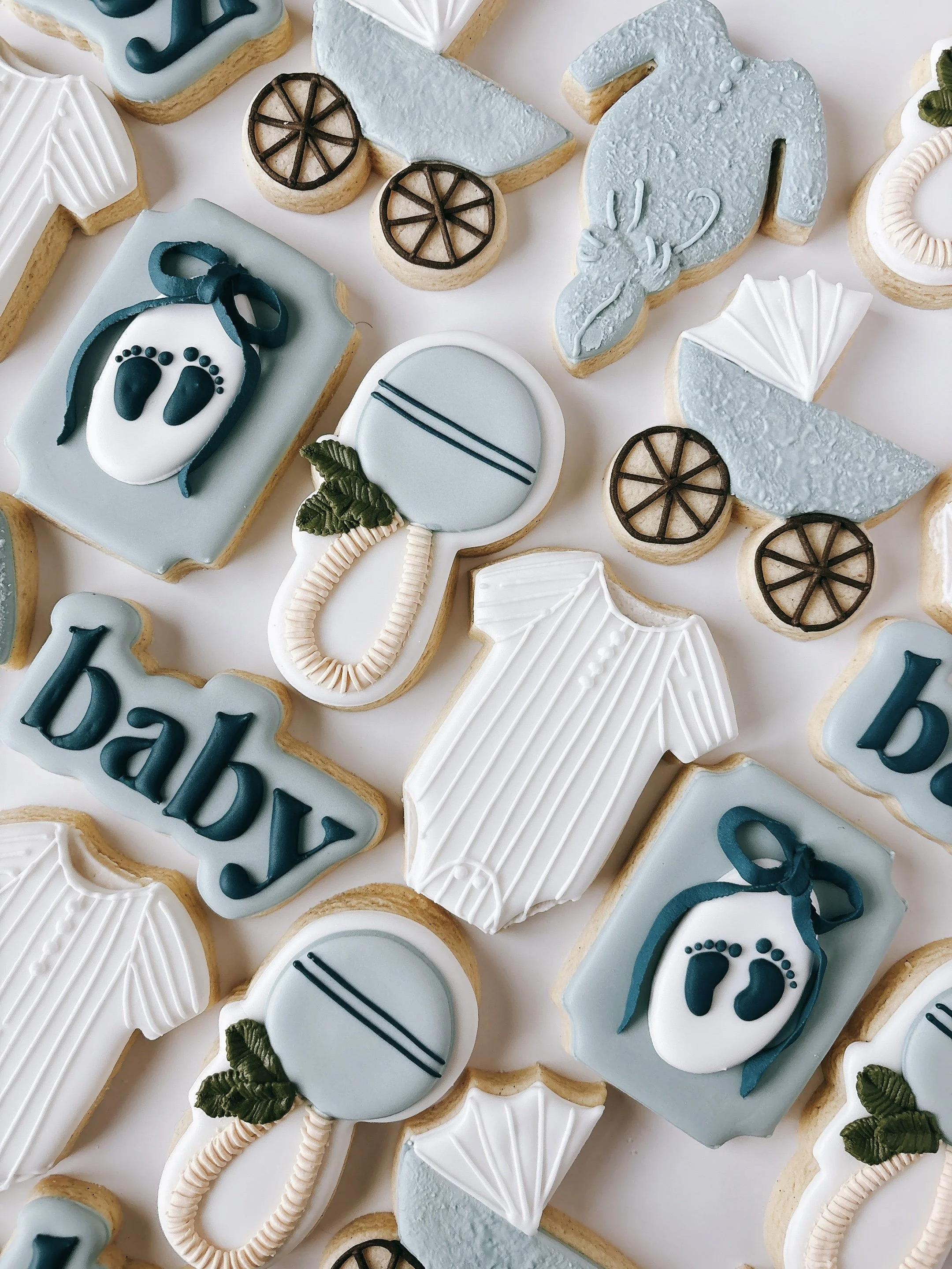 Assortment of baby shower themed cookies with blue, white, and brown icing, decorated as pacifiers, baby booties, strollers, and the word "baby".