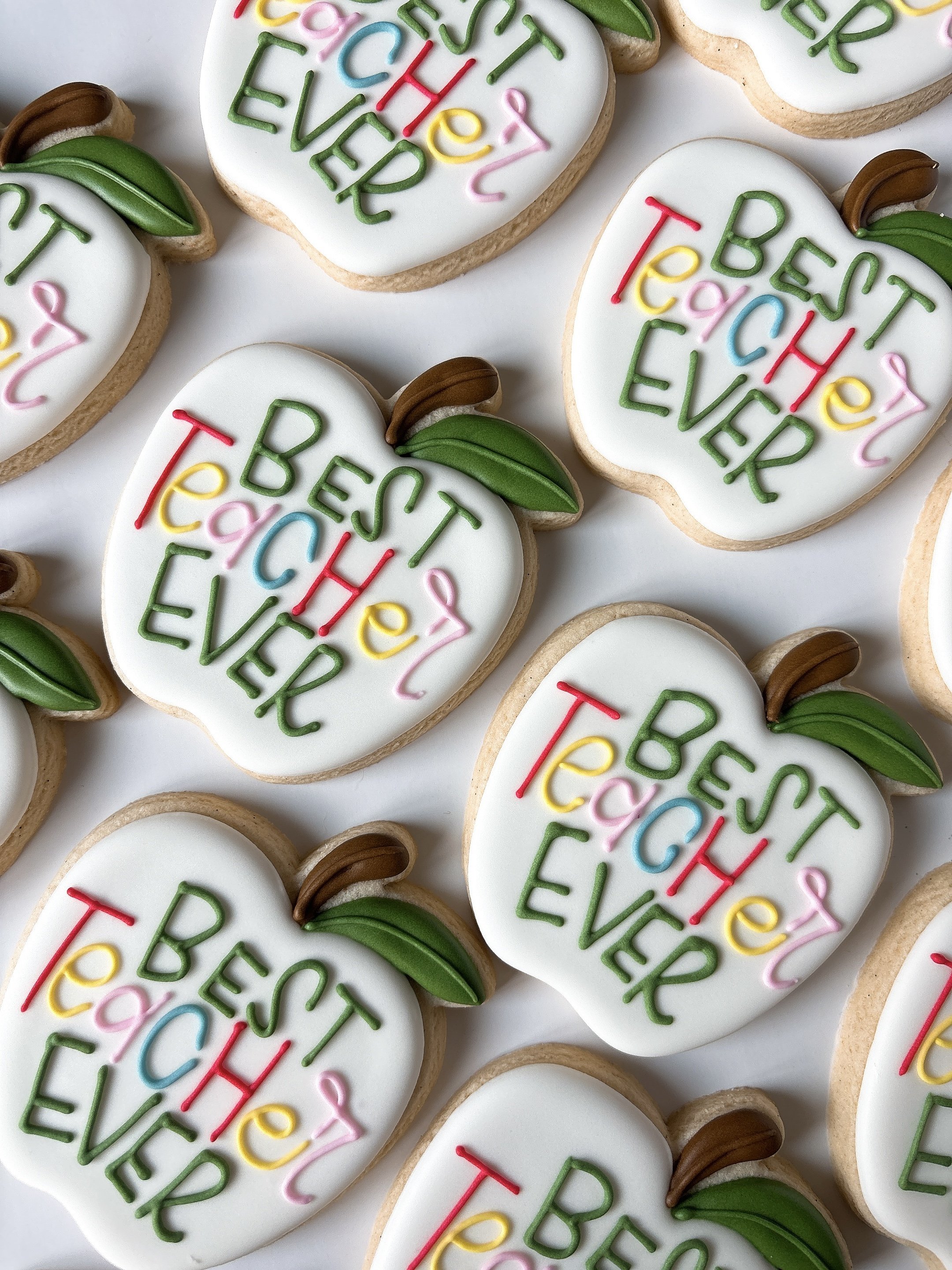 Heart-shaped cookies decorated with white icing, green, red, pink, yellow, and blue icing words reading "Best Teacher Ever" with green leaves and brown stem decorations.