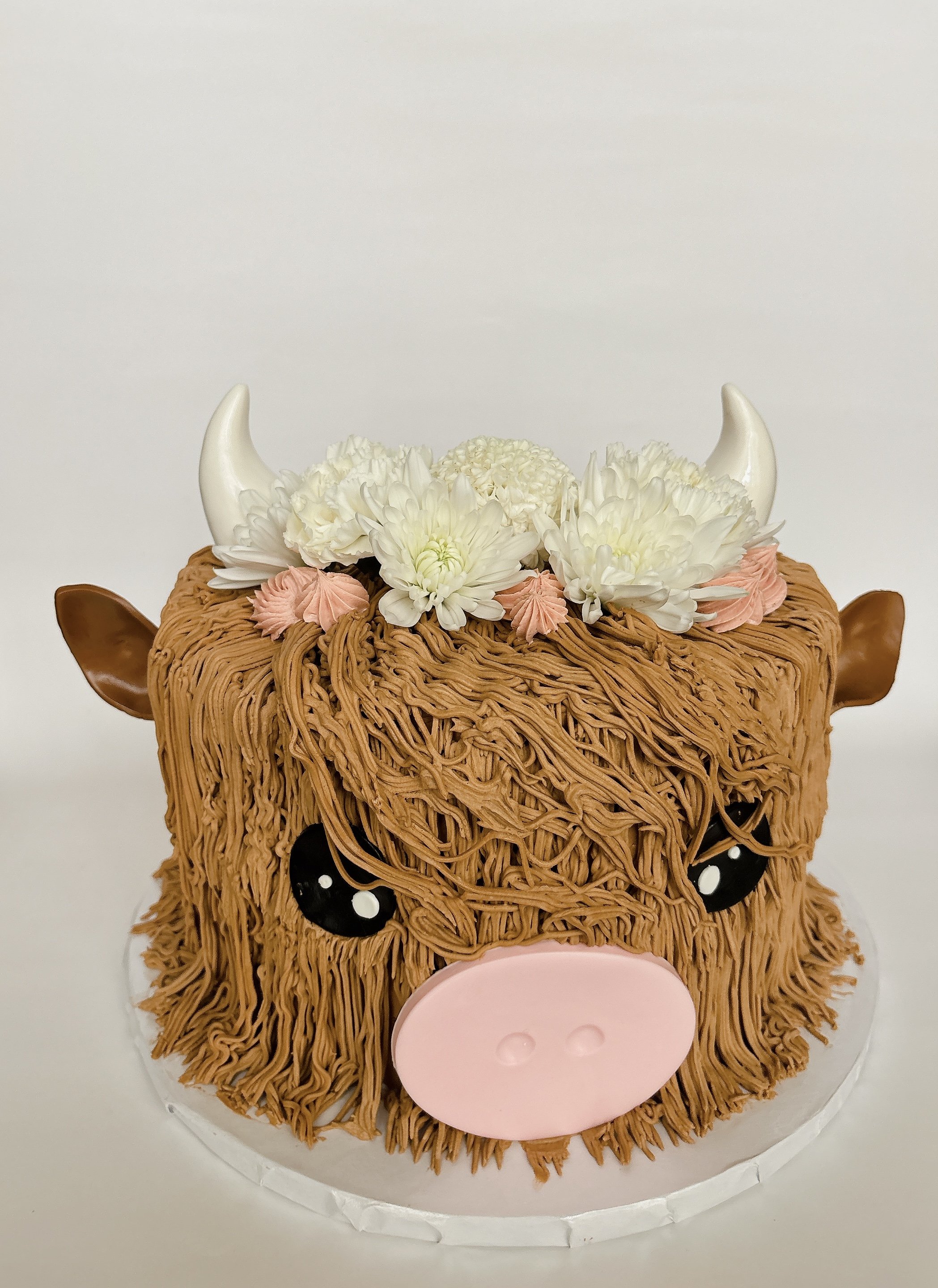 Cake decorated as a cow with chocolate fur, white horns, black and white eyes, pink nose, and floral flowers on top.