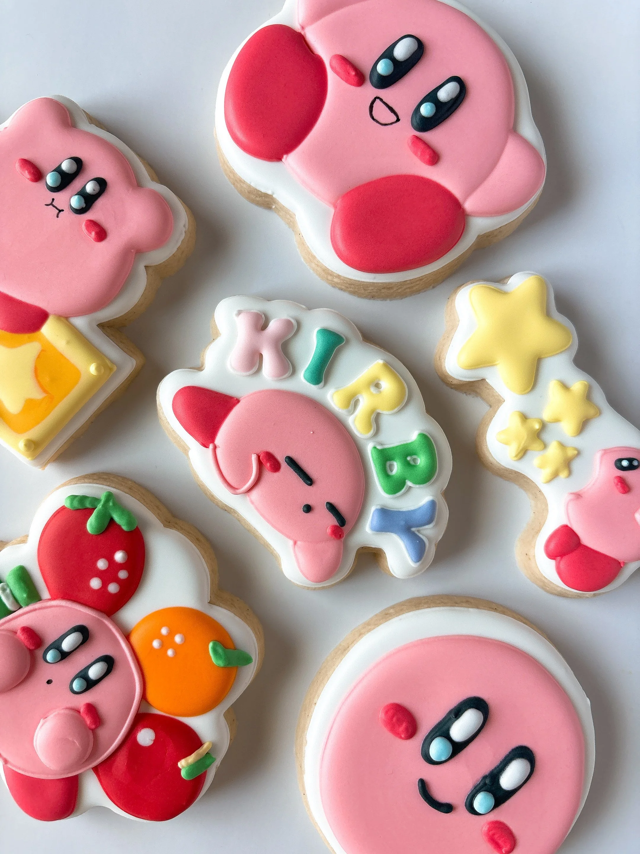 Colorful decorated cookies in various shapes, featuring pink, yellow, orange, and red icing with characters and patterns, including a cute pink star and a cookie with the words 'KYS' and hearts.