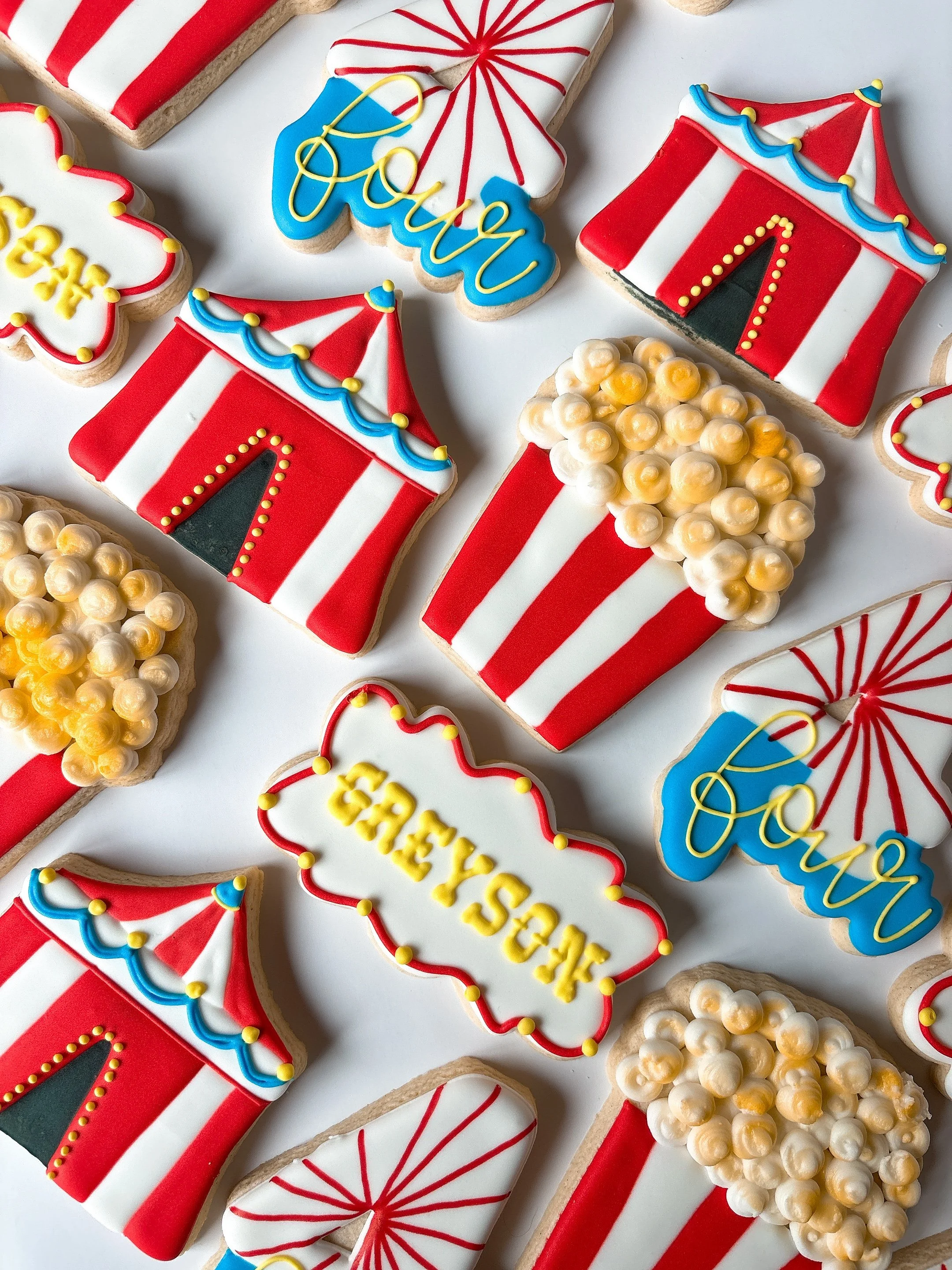 Decorated cookies themed for a circus and celebration with red, white, blue, and yellow icing, featuring tents, ferris wheel, and messages like "Party" and "Cheers."