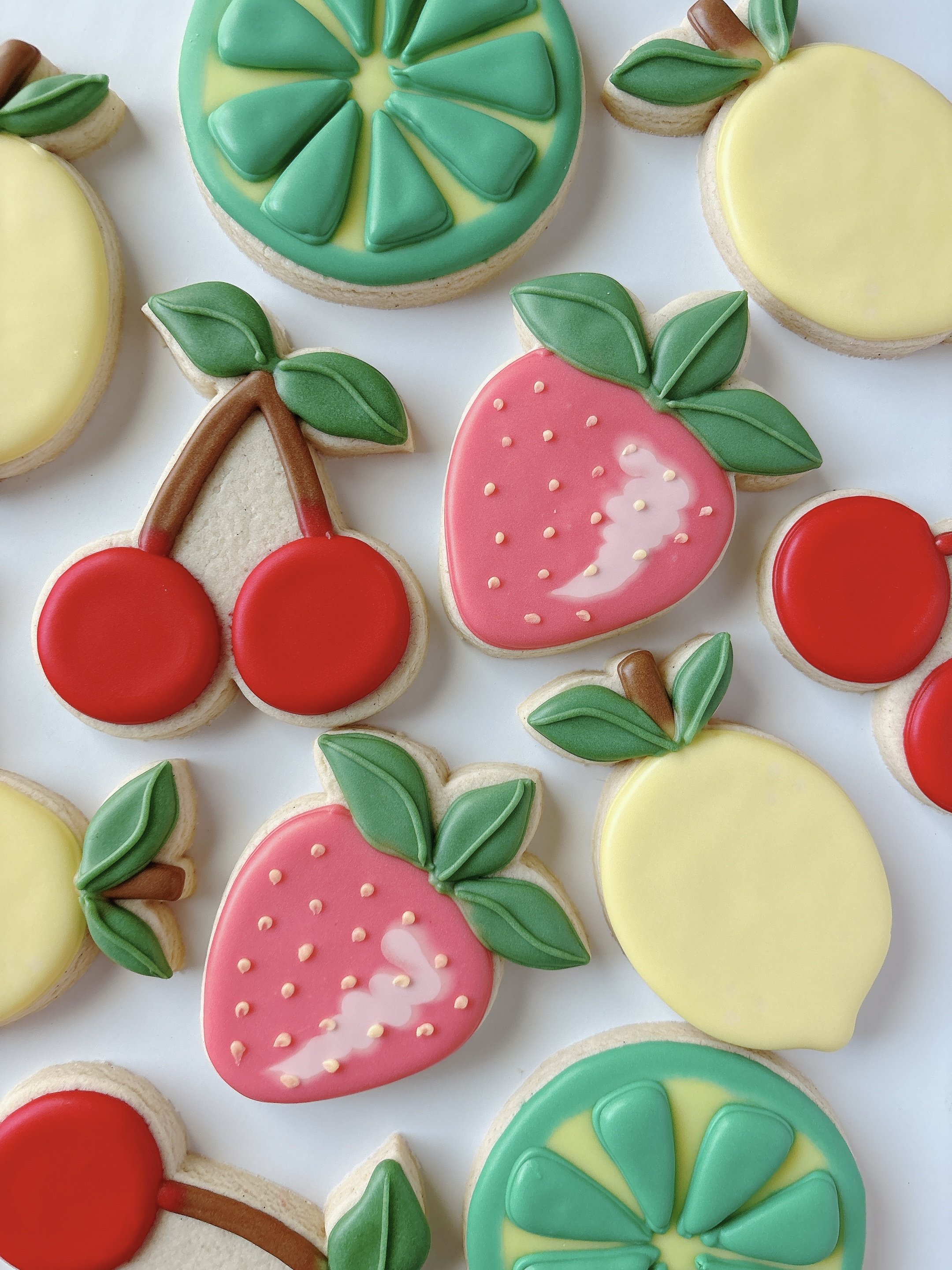 Colorfully decorated sugar cookies shaped like slices of citrus fruit and cherries, with green leaves and vibrant icing.