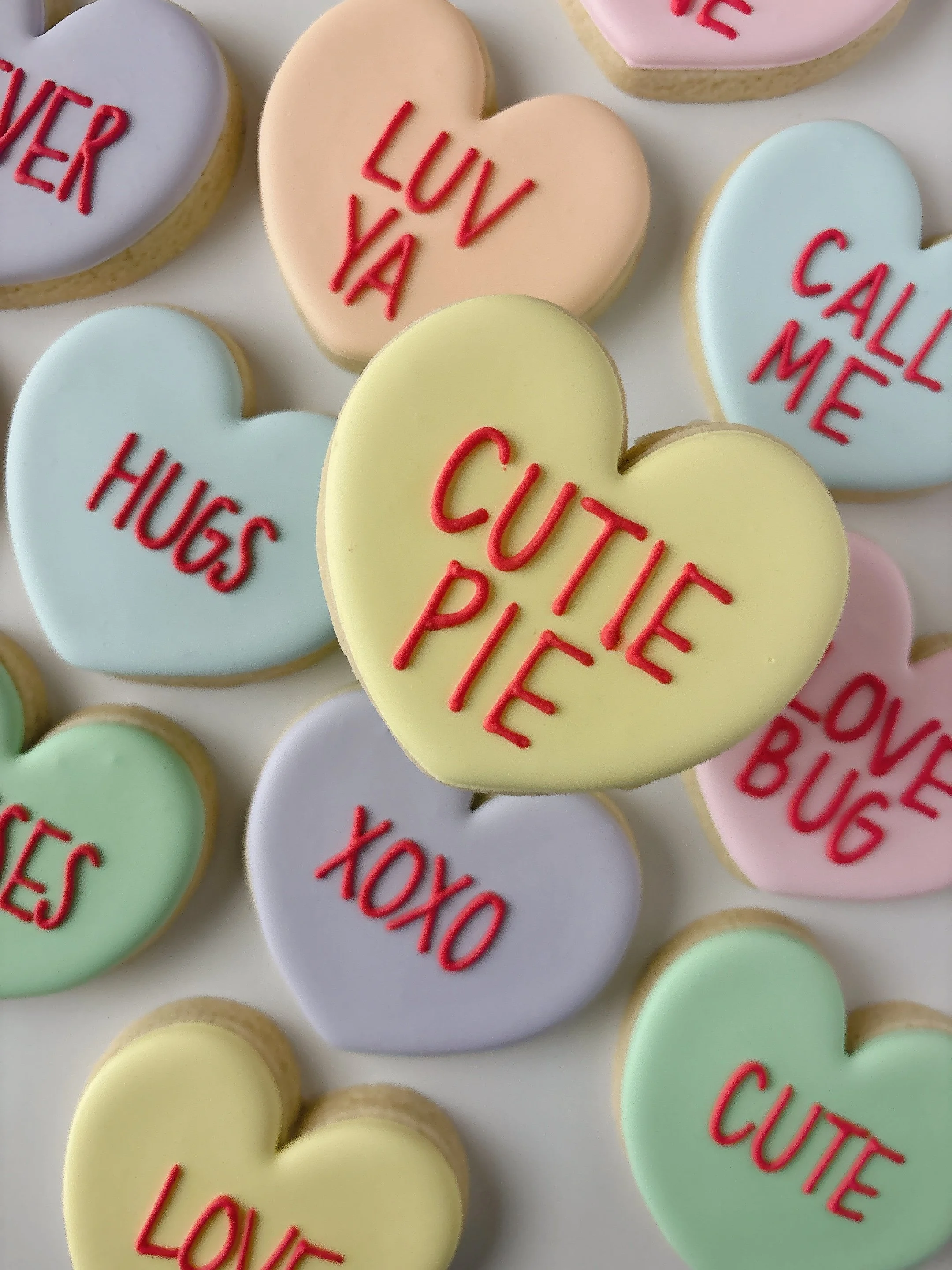 Heart-shaped cookies with pastel icing in colors like yellow, pink, blue, and purple, each decorated with red icing words such as "LOVE," "HUGS," "CUTE," "CALL ME," and "YOU."