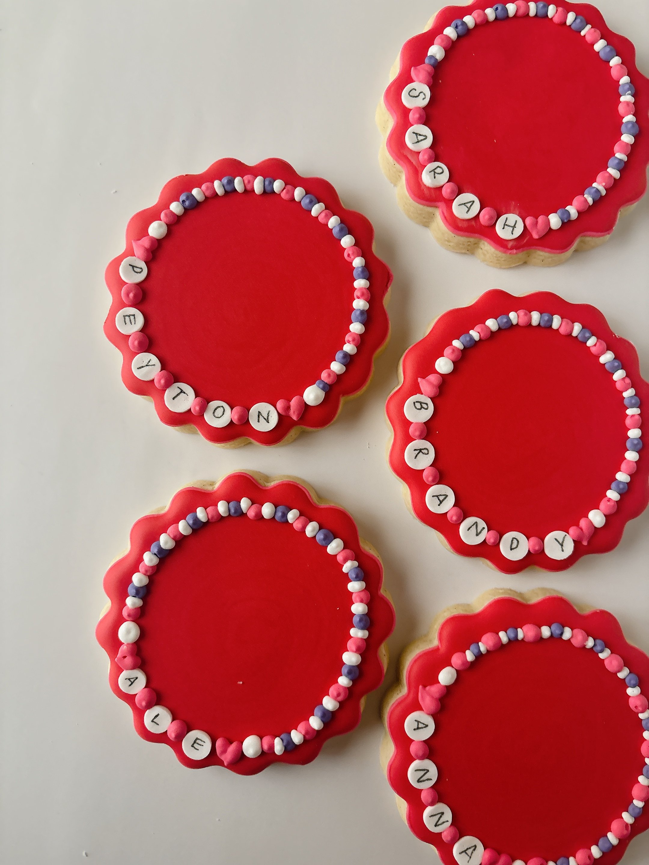 Decorated cookies with red icing and a border of pink, white, and purple sugar beads, each cookie featuring a letters that spell out various names.
