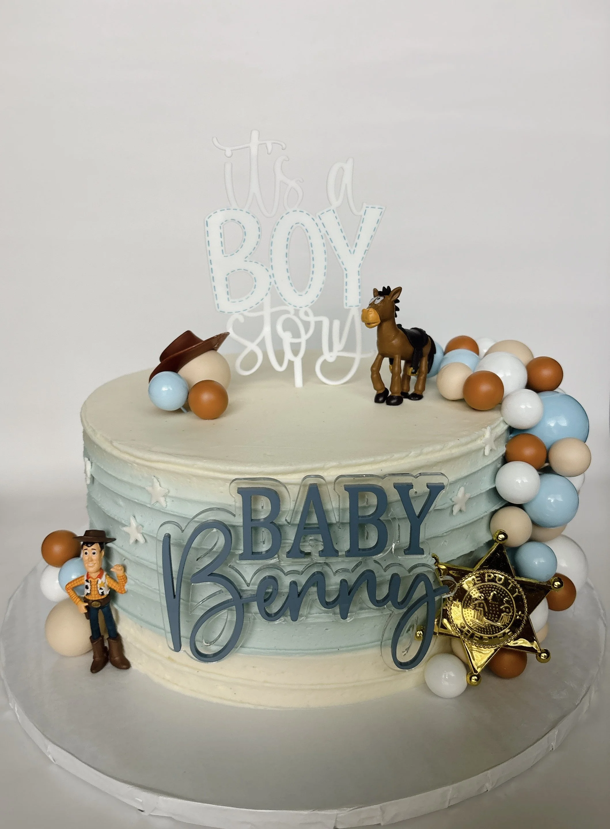 A cake decorated for a baby boy gender reveal. The cake has a topper that reads 'It's a Boy' and features Toy Story characters Woody and Bullseye. The cake is adorned with multicolored candy-like balls and a gold badge.
