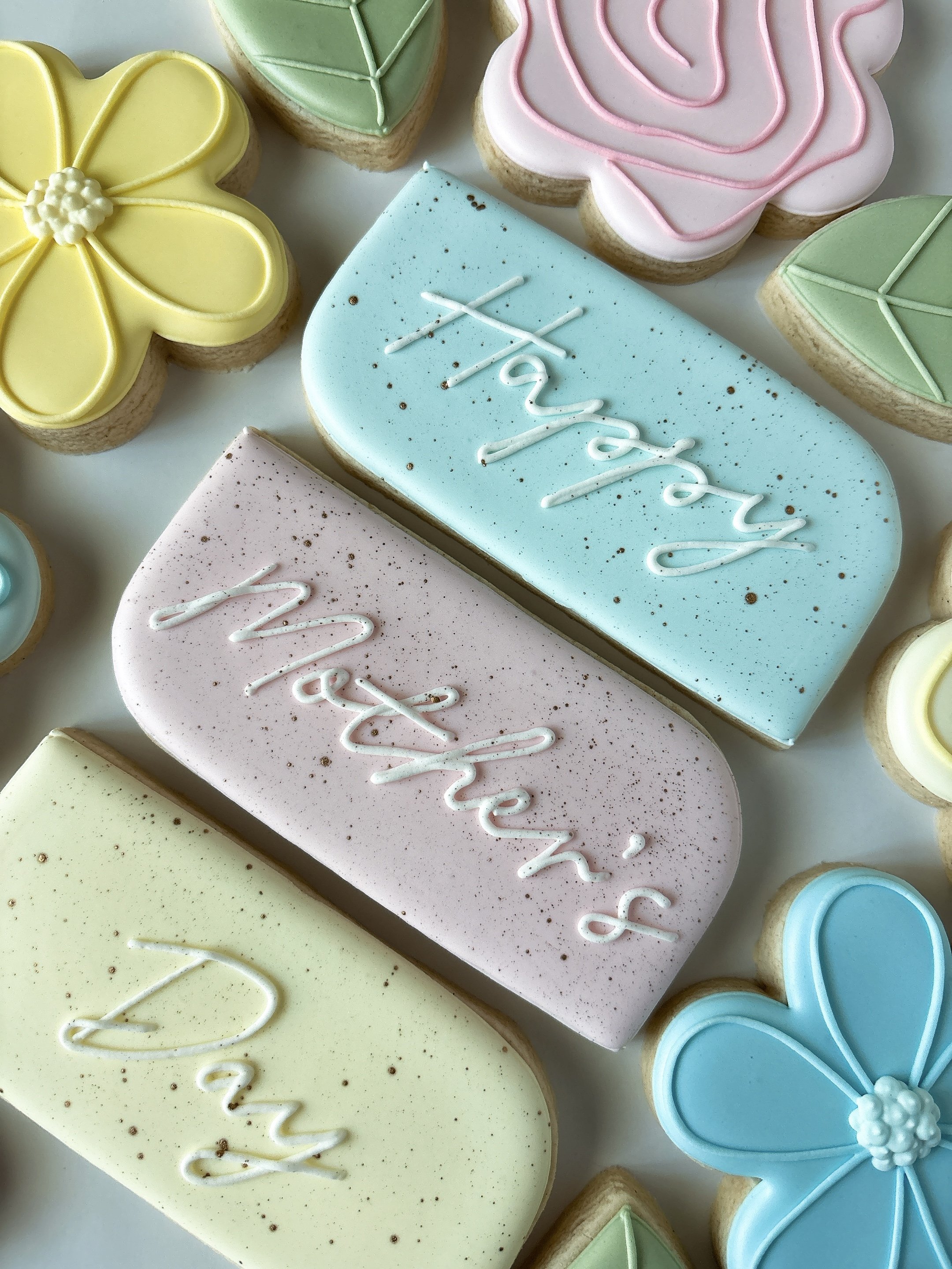 Colorful decorated cookies with pastel icing in the shape of flowers, leaves, and a butterfly, with some cookies having the word 'mother' written on them.