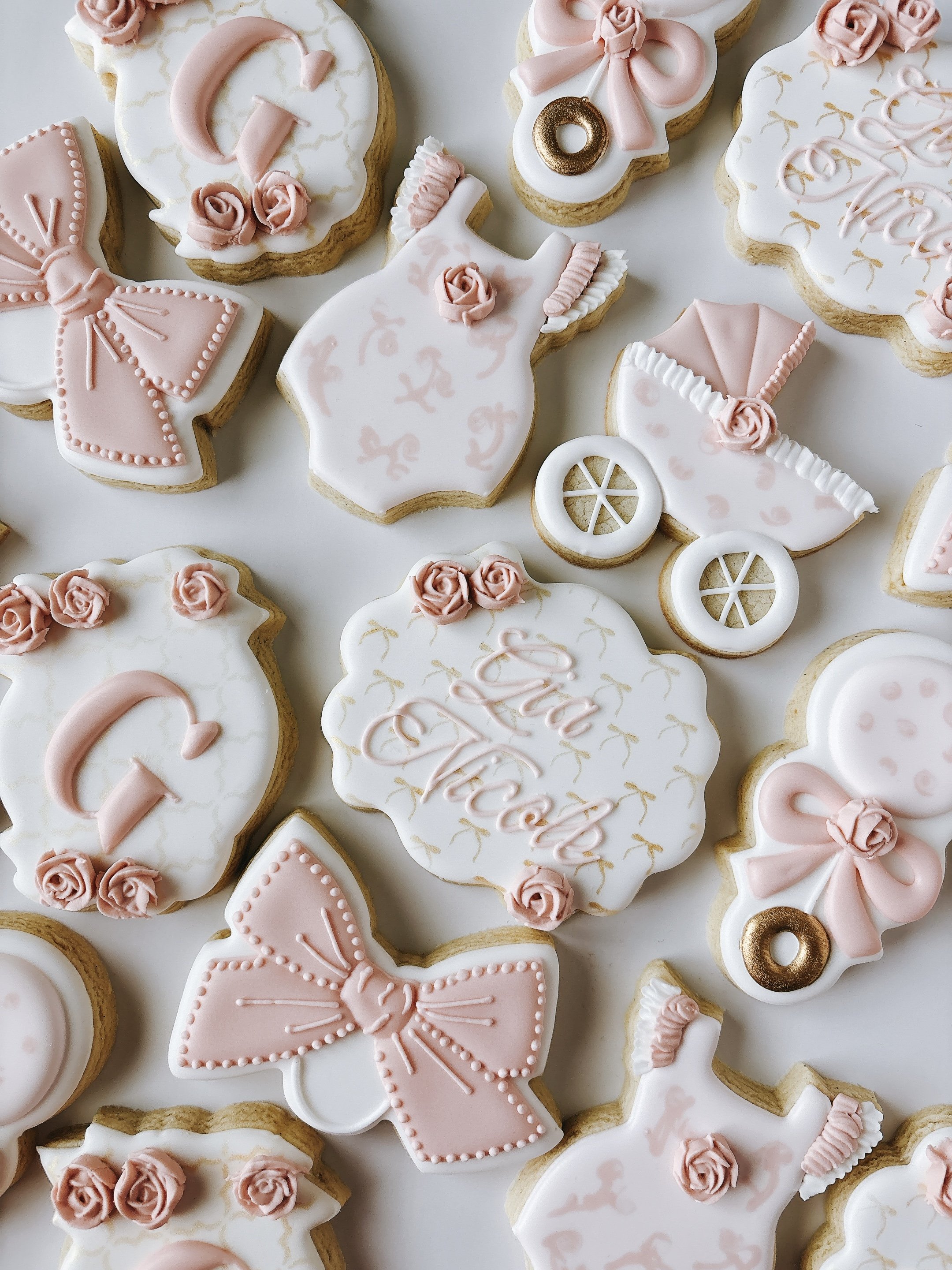 A variety of decorated cookies with pink and white icing, including a rattle, a baby carriage, bows, and girly onesies.