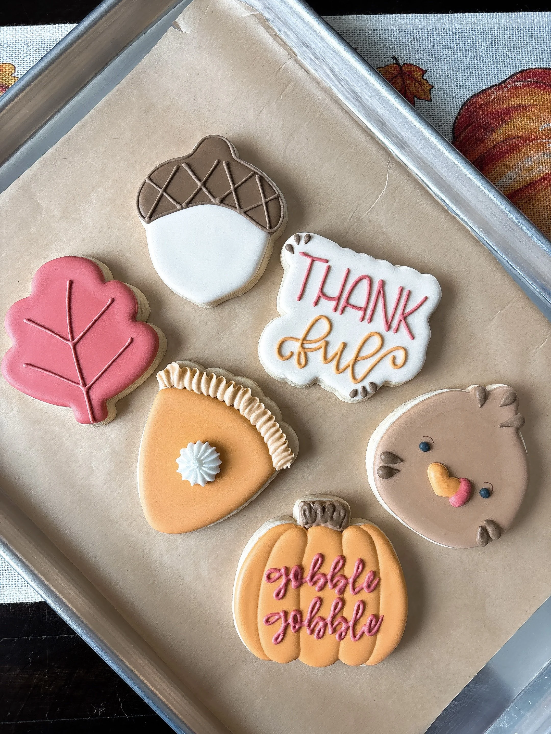 Decorated fall-themed cookies on a baking sheet, including a pink leaf, a pumpkin with pink writing, a white cookie with a thank you message, and a bear face.