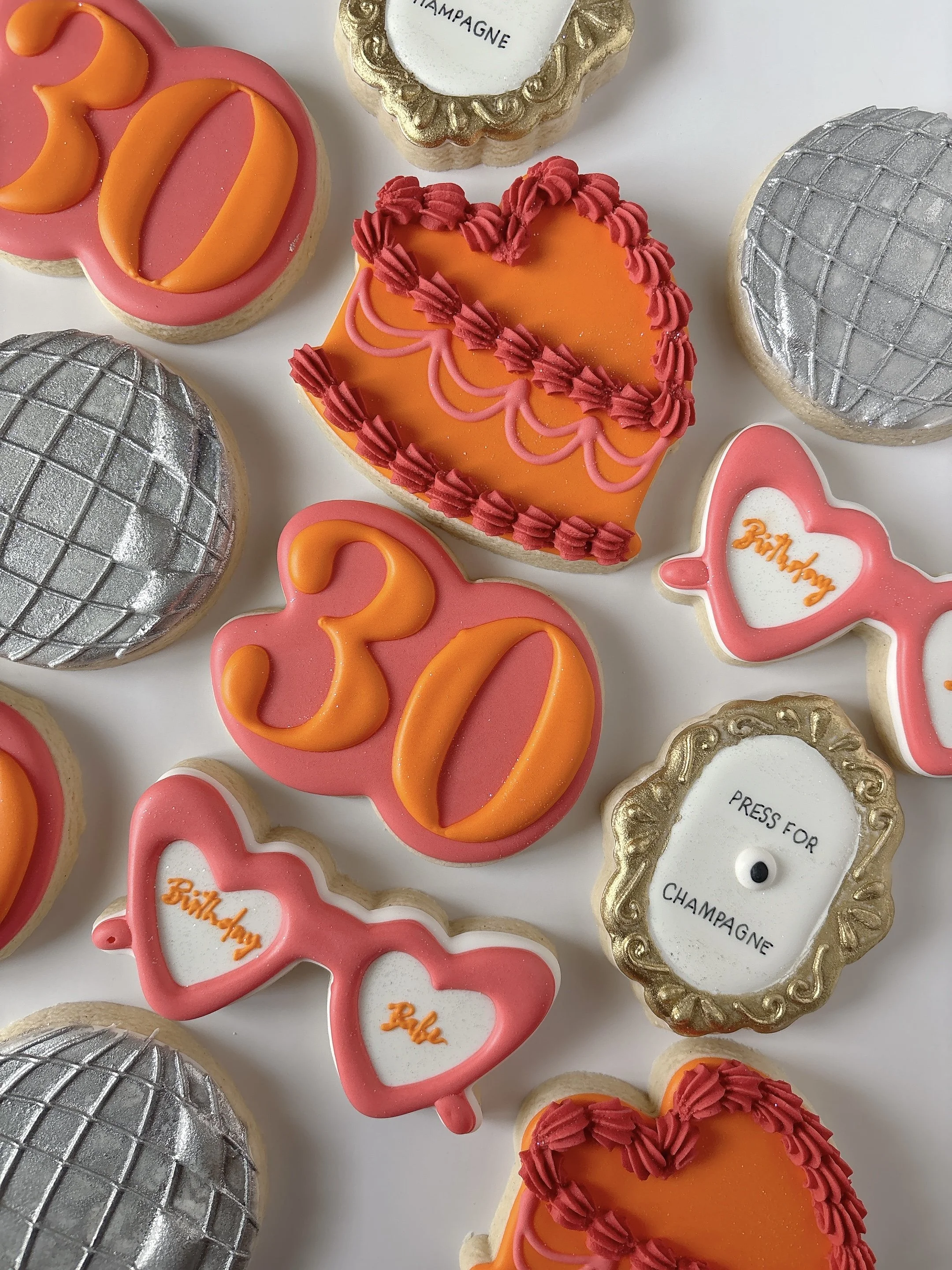 Decorative cookies shaped like hearts with pink, orange, and silver icing, some with the number '32' and others with the words 'Birthday' and 'Press for Champagne' on them.