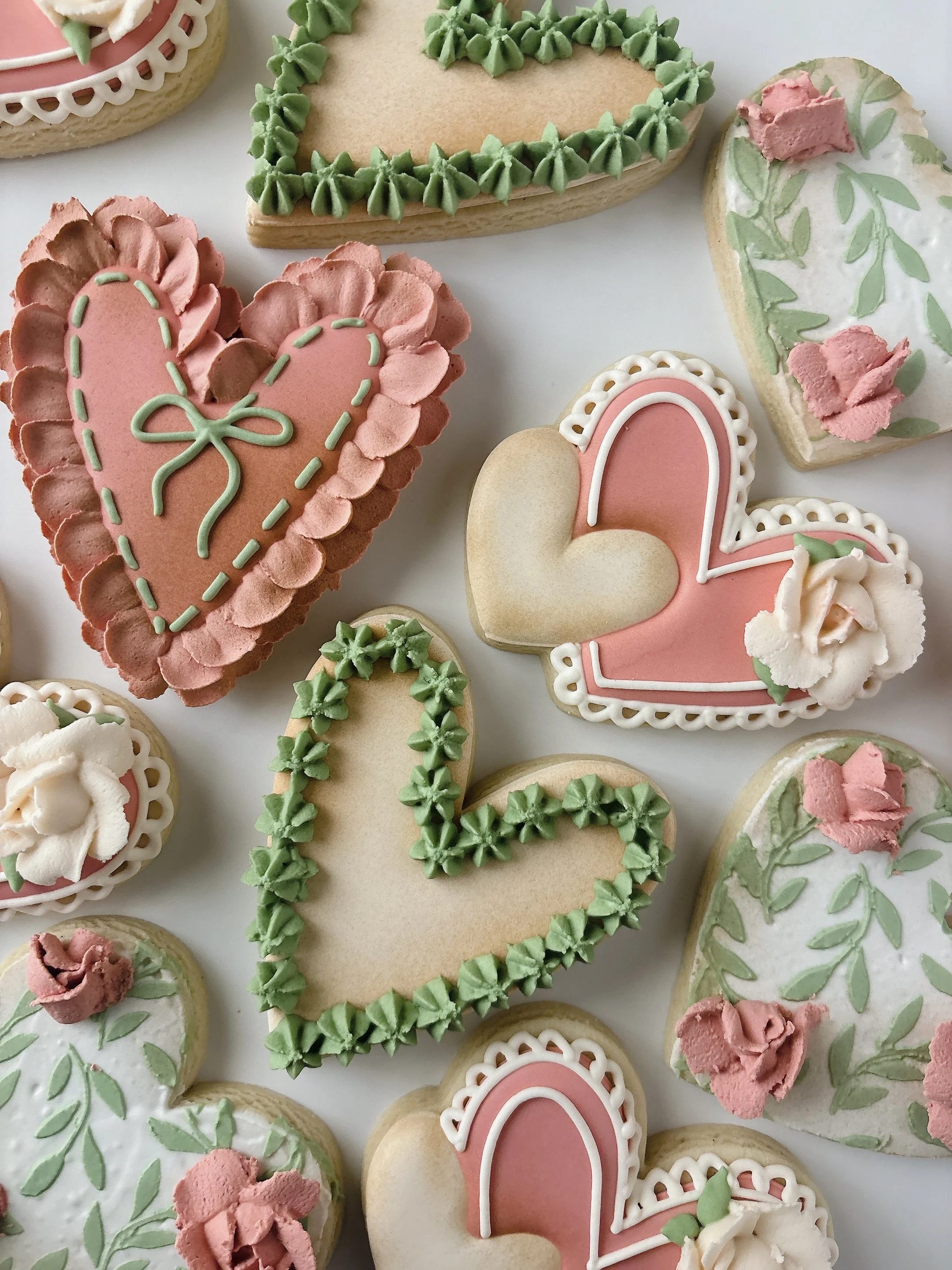 Assorted decorated cookies with pink, green, and white icing in various heart, flower, and abstract shapes on a white background.