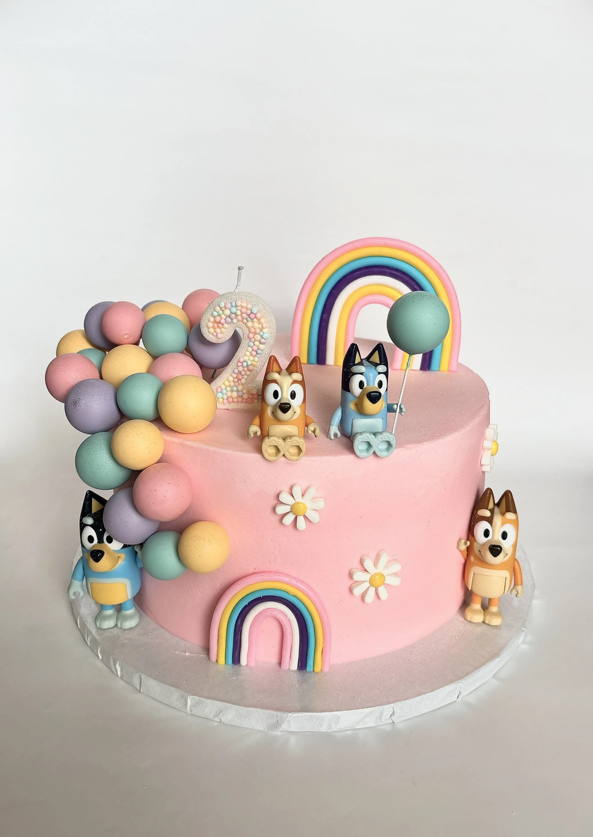 Pink birthday cake decorated with pastel-colored balloon beads, toy animal figurines, a rainbow, flower decorations, and a large number 2