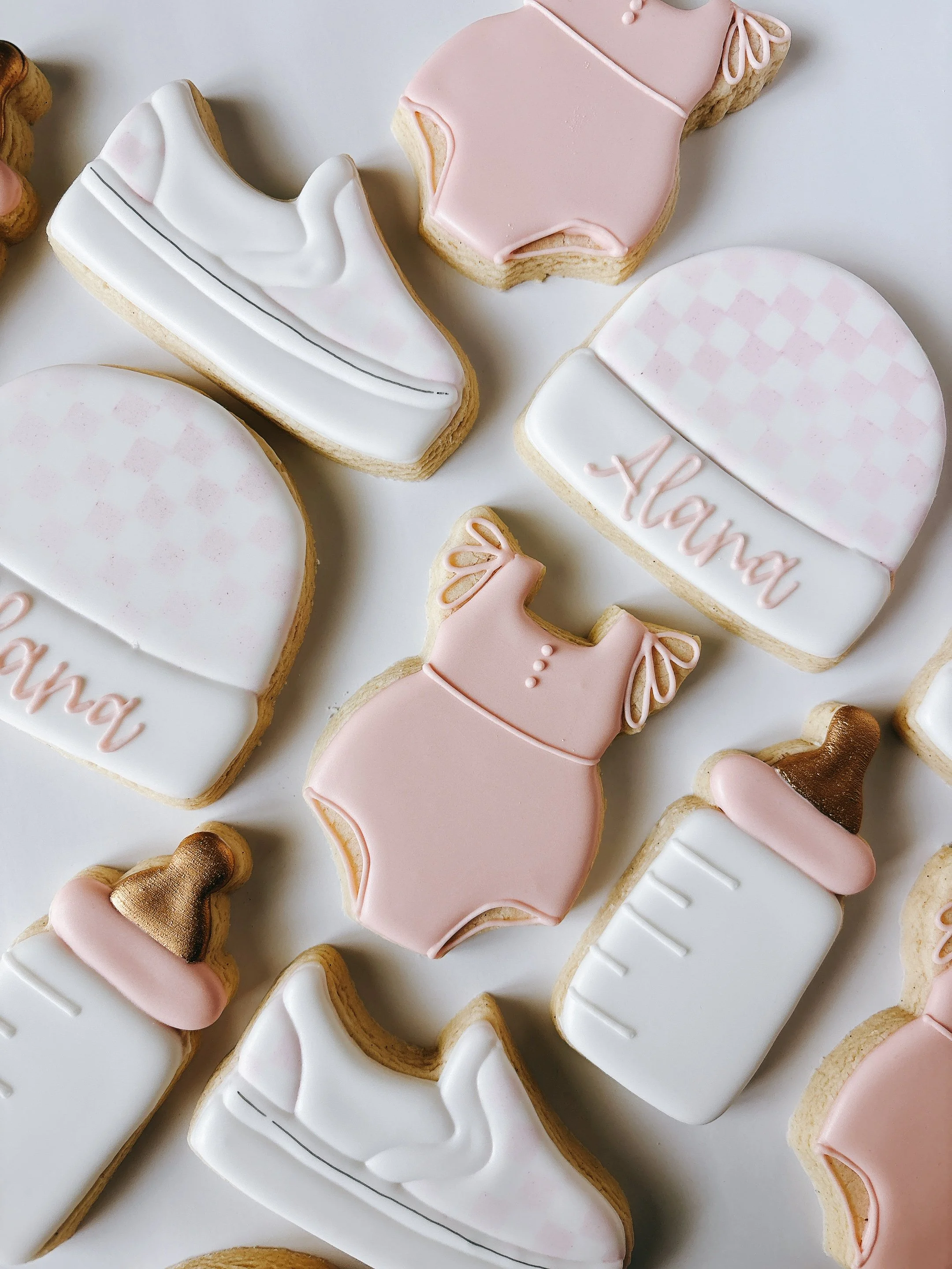 Assorted baby shower cookies decorated with pink, white, and gold icing, shaped like baby clothes, bottles, and accessories, with some cookies featuring the word "Mom".