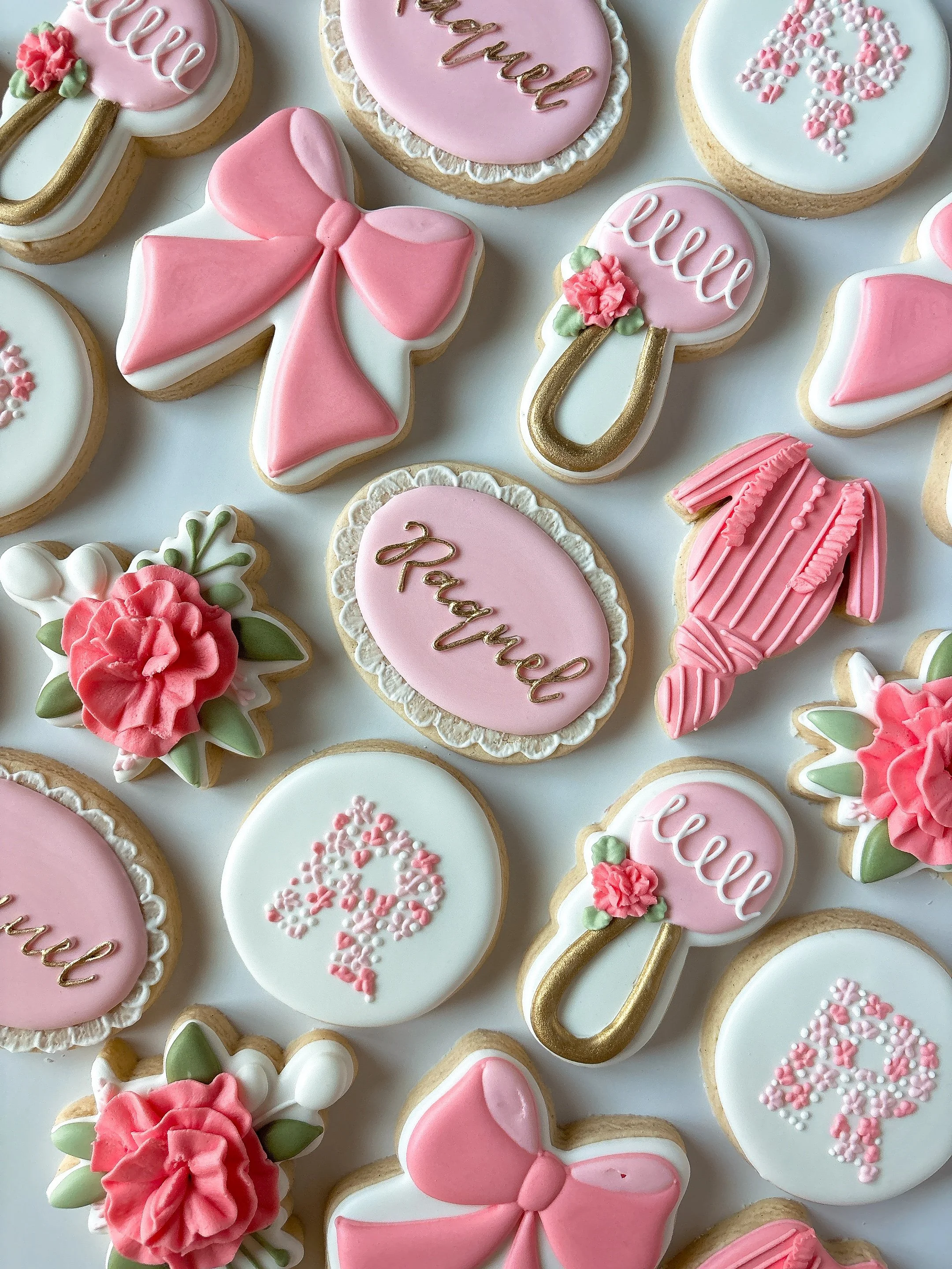 Decorated cookies with pink, white, and gold icing, featuring hearts, flowers, a bow, and phrases like 'hello' and 'prayer' in elegant script.