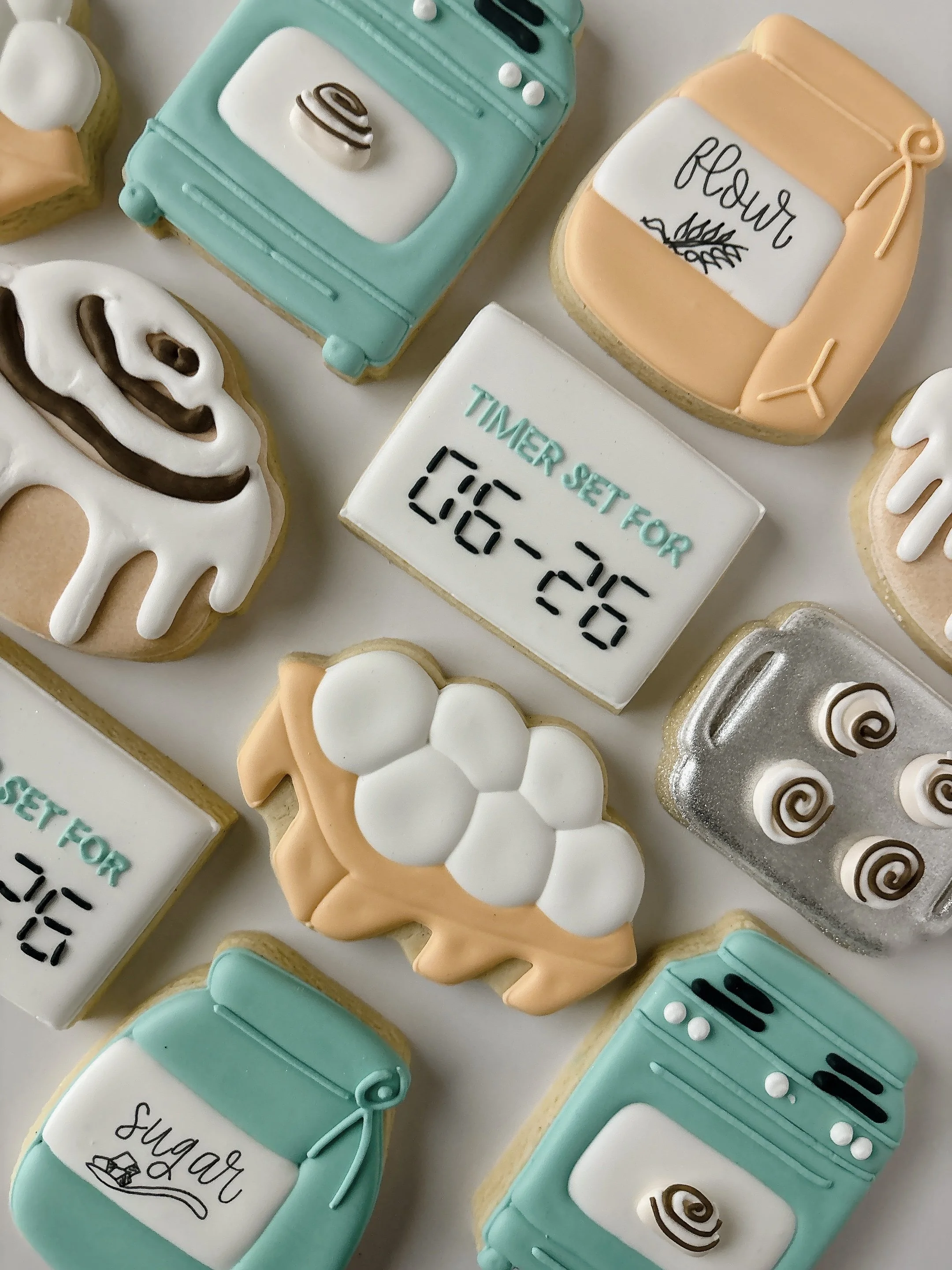 Decorated cookies shaped like kitchen appliances such as an oven, a sugar bag, a flour bag, and eggs with pastel colors and writing.