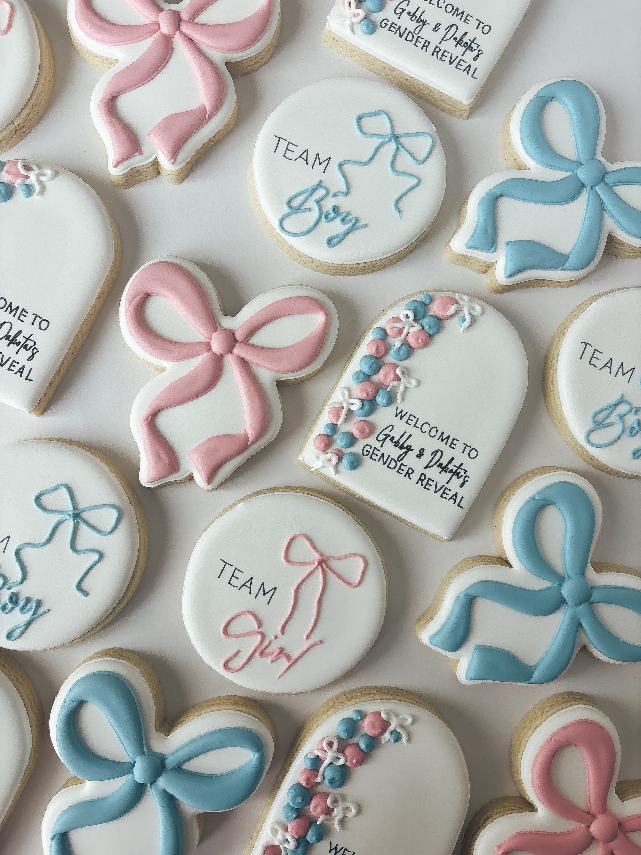 Decorated sugar cookies for a gender reveal celebration. The cookies are in pink and blue, including bows, ribbons, and solid colors, with some featuring writing that says 'Team Girl' and 'Team Boy' with additional greetings.