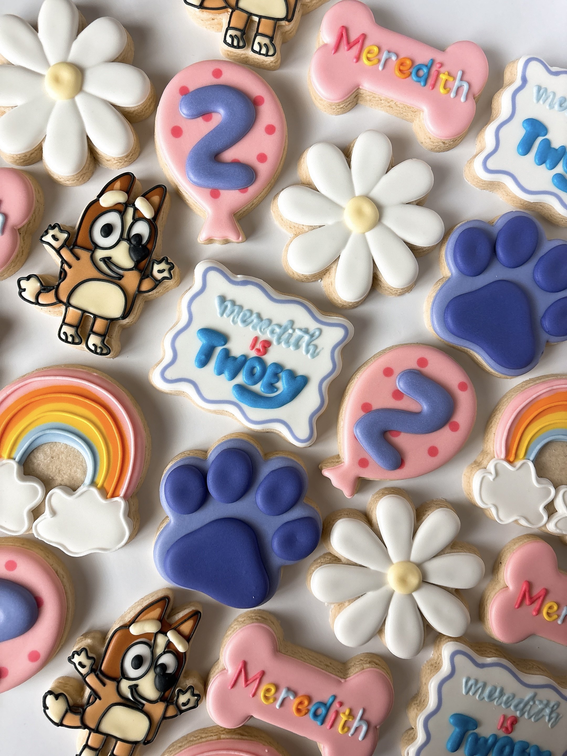 Decorative cookies with various designs including a white daisy flower, a cartoon dog with glasses, a pink bone-shaped cookie with multicolored text, a pink balloon-shaped cookie with the number 2, a cloud-shaped cookie with the message 'Meredith is 