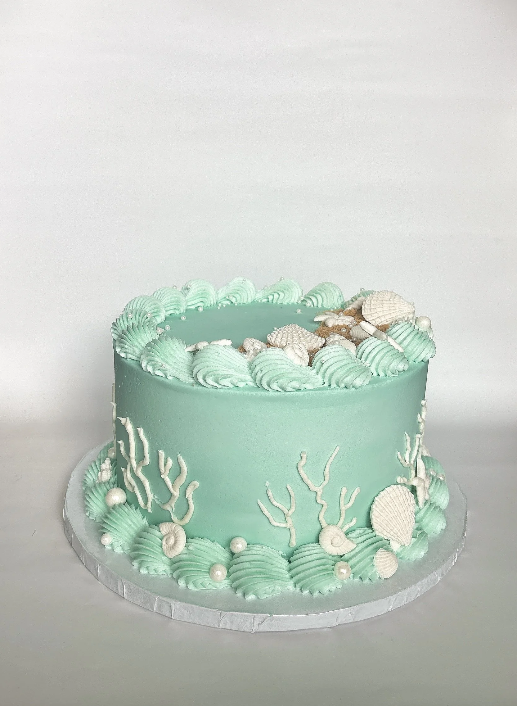 A single-tiered ocean-themed cake decorated with turquoise and white icing, featuring seashell and coral-shaped designs, with white pearl-like accents.
