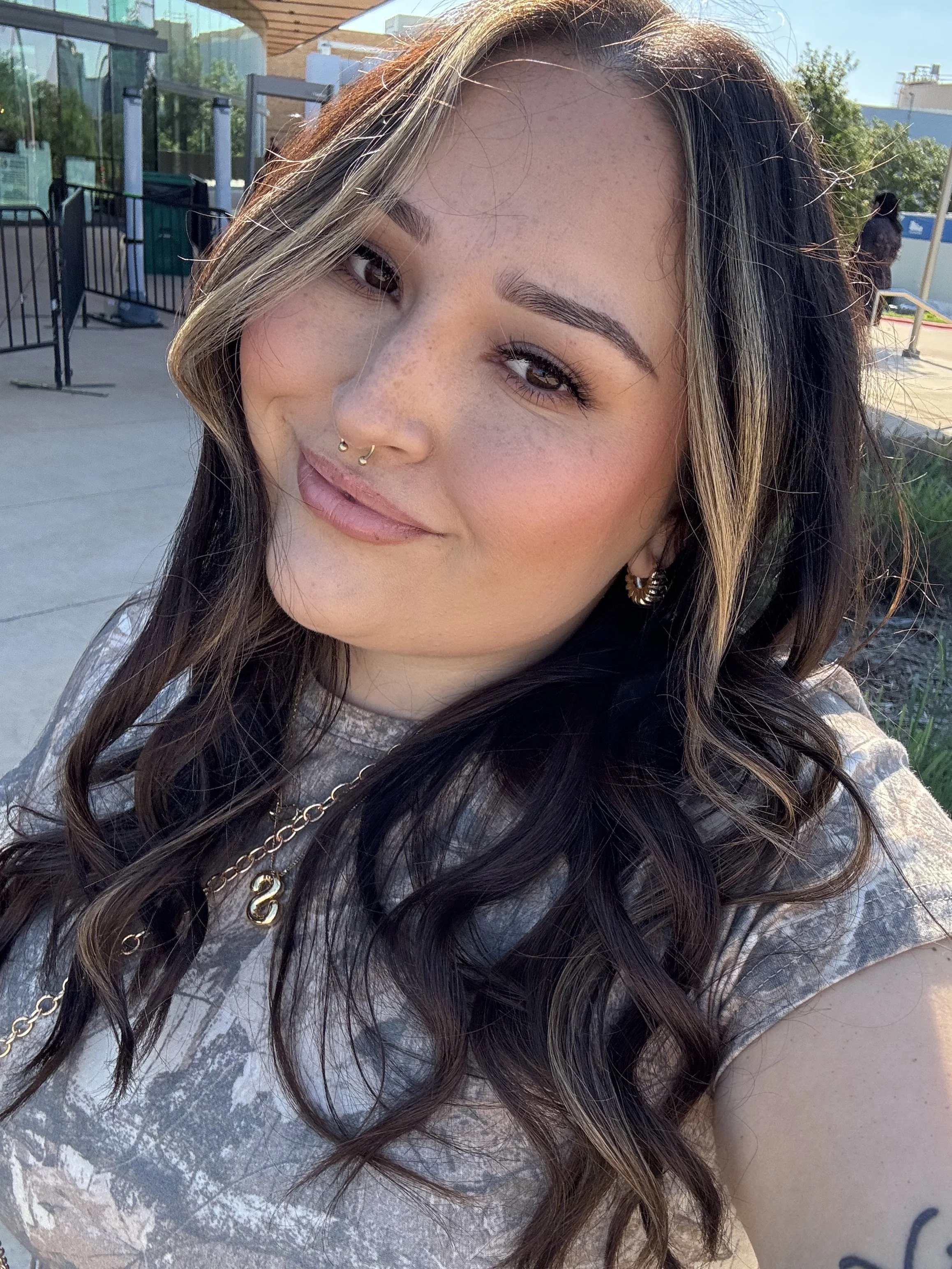 A woman with long dark hair and light highlights, smiling slightly, outdoors on a sunny day. She has physical features such as freckles, a septum piercing, and is wearing earrings and layered necklaces. Part of her gray and white tie-dye shirt is visible.