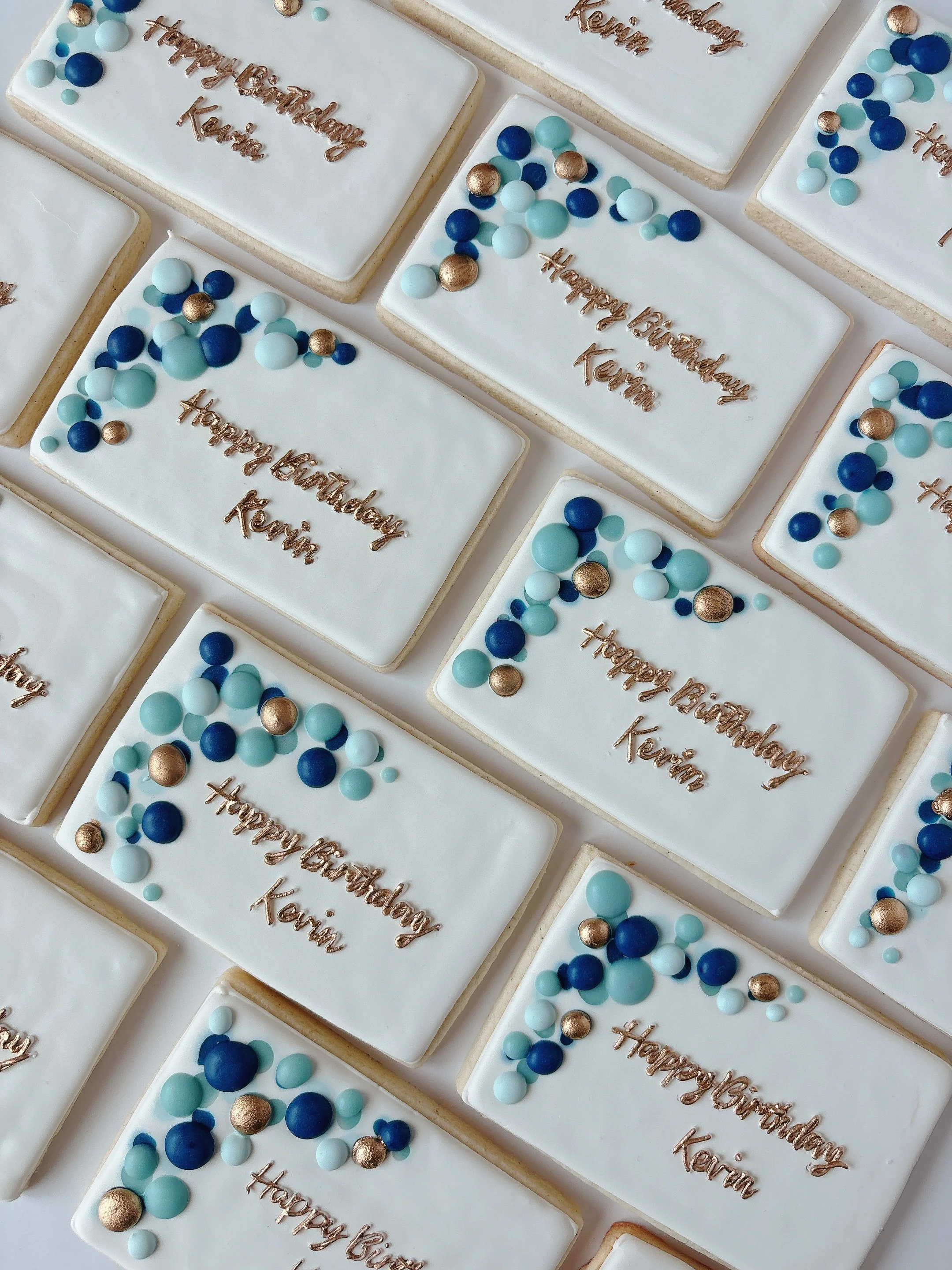 Several rectangular cookies decorated with white icing, blue and gold spherical candies, and a message written in gold script that reads 'Happy Birthday Kavina'.