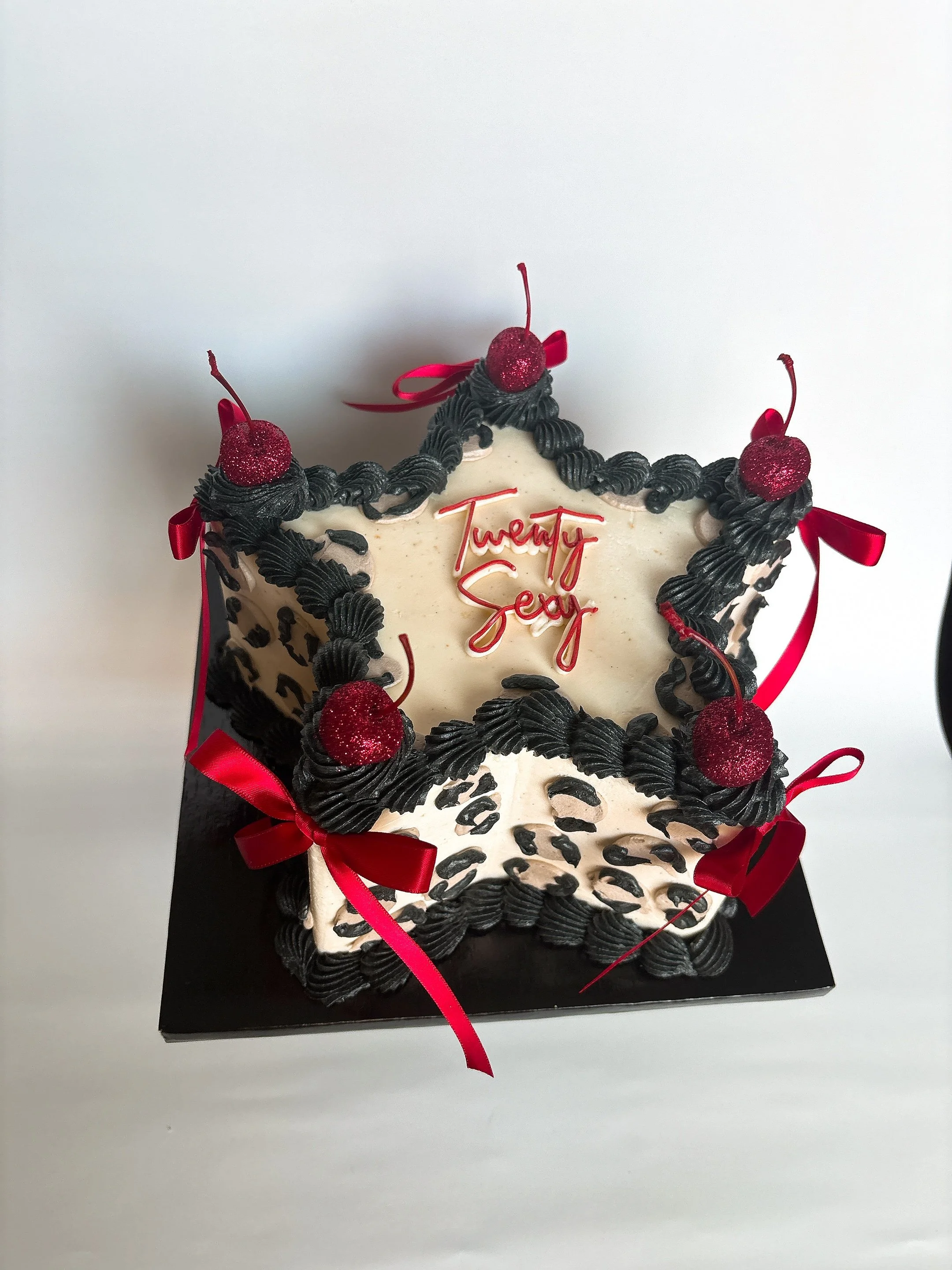 A white birthday cake decorated with black icing, red cherries with glitter, red ribbons, and the words "Twenty Sexy" written in red on top.