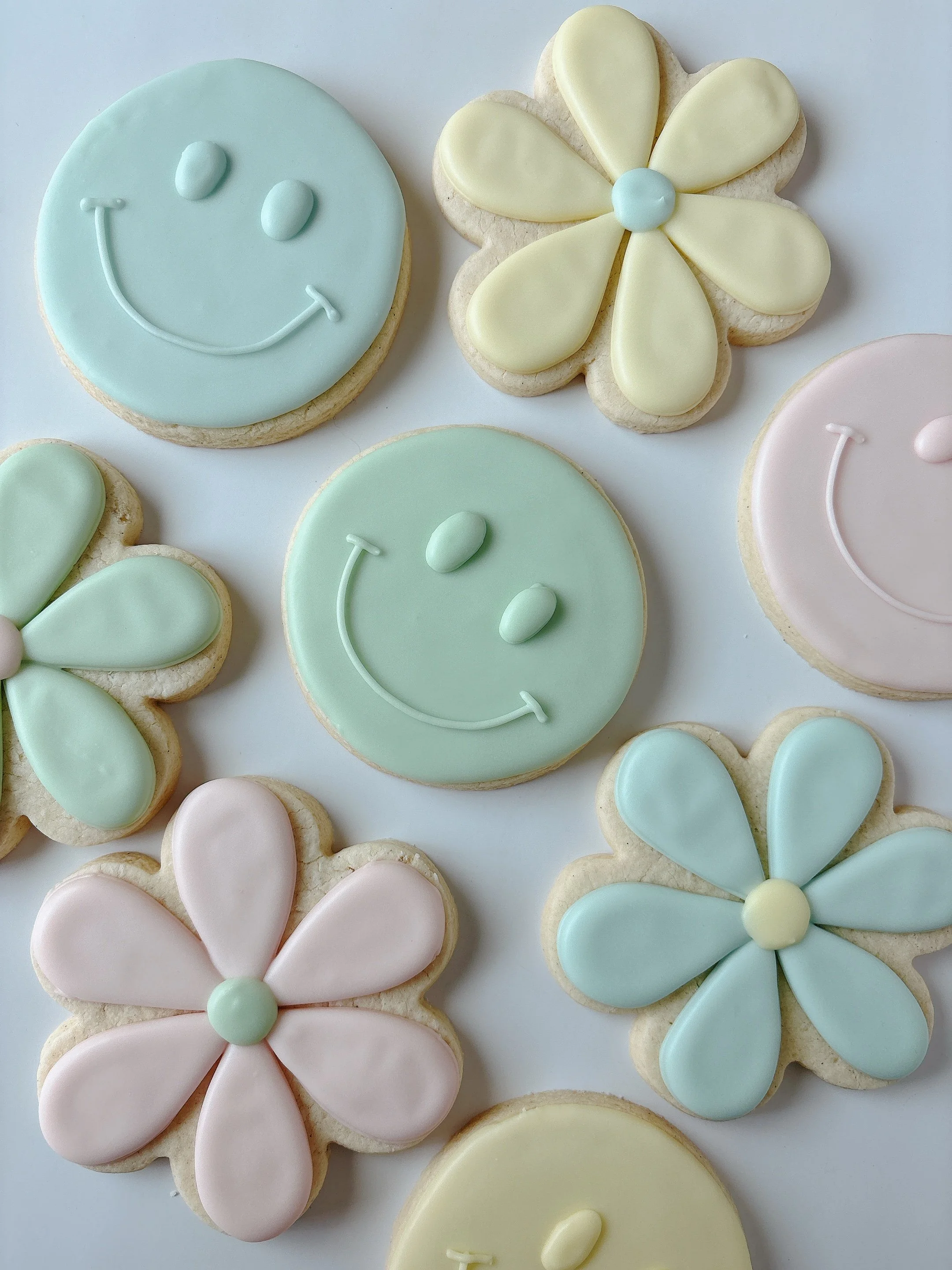 Cookies decorated with pastel icing in the shapes of smiley faces, flowers, hearts, and butterflies.
